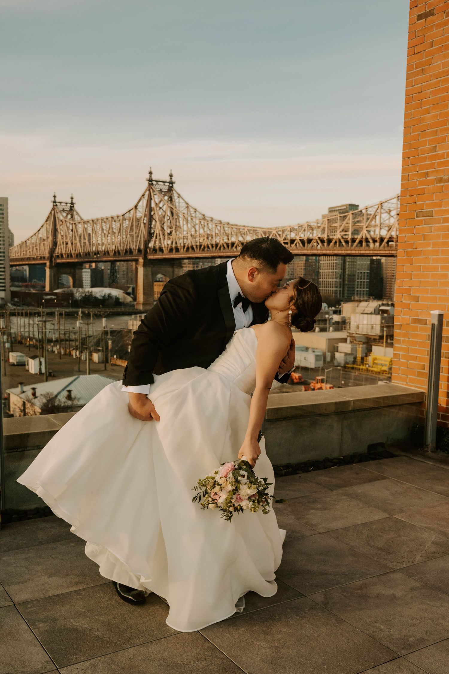 Romantic wedding couple shares a dip kiss on rooftop with Queensboro Bridge in New York City backdrop at sunset.