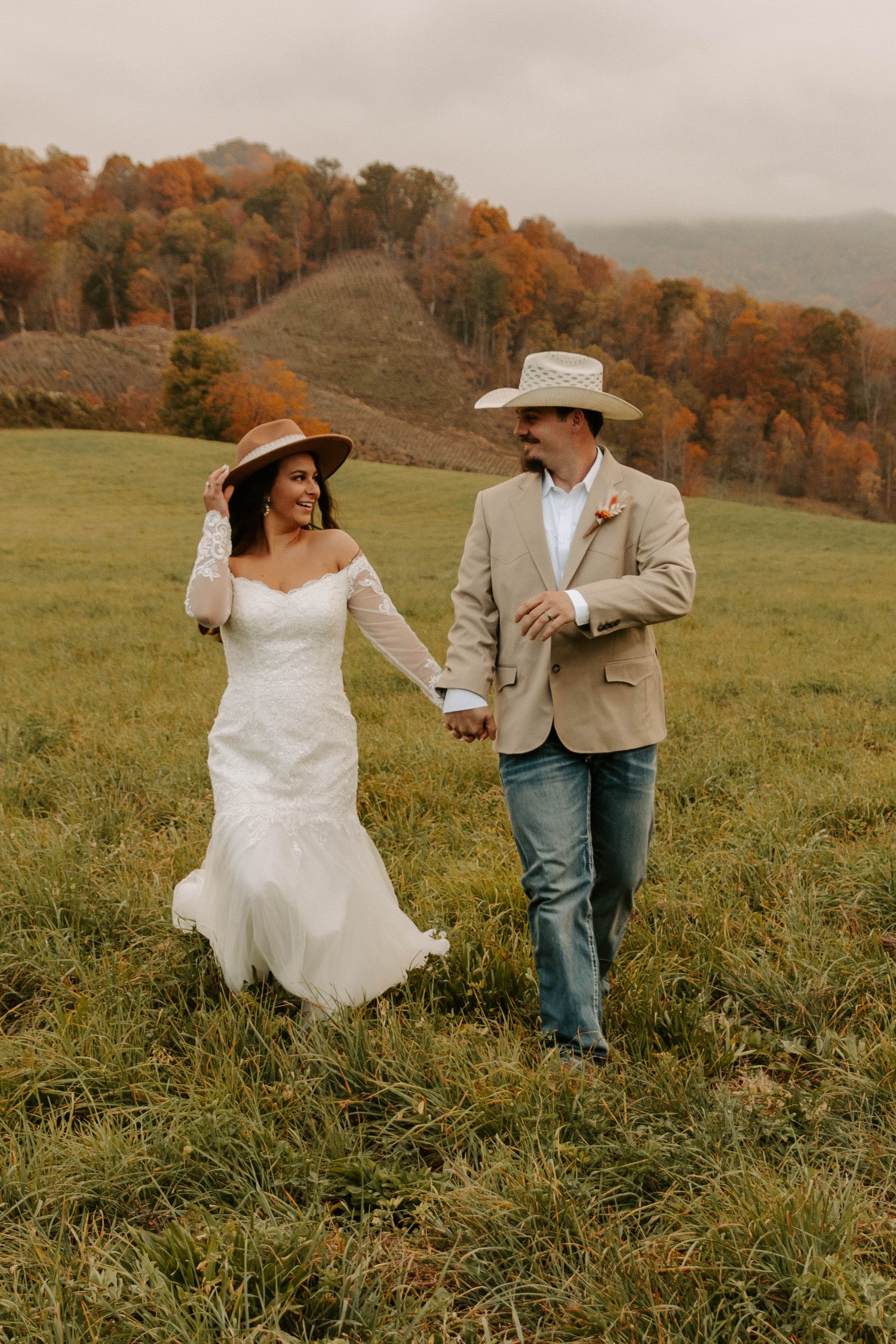 A couple in western-style attire walks hand in hand through an autumn meadow with colorful fall foliage on hillside behind them.