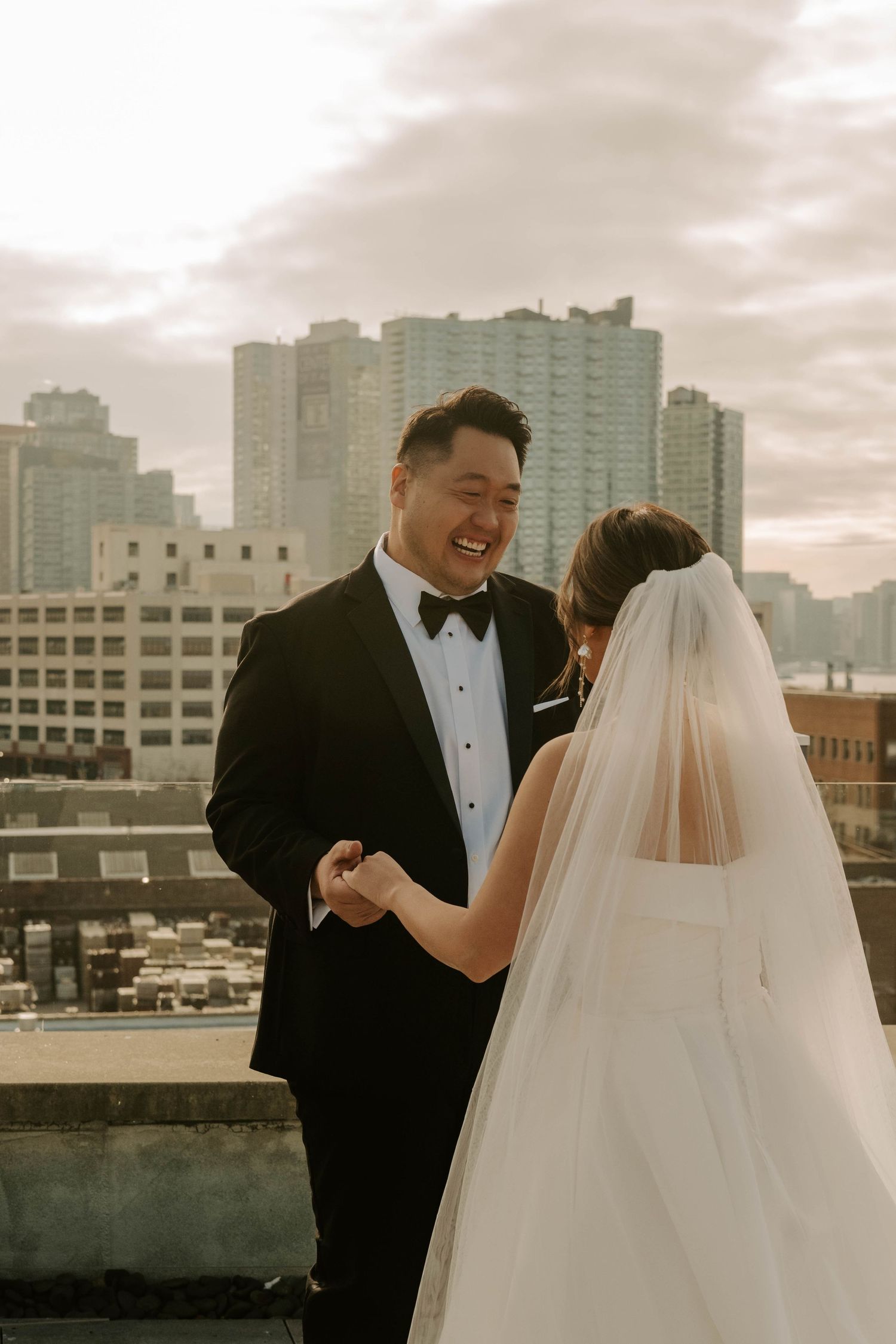 A bride and groom share a joyful moment on a rooftop with city buildings in the background.