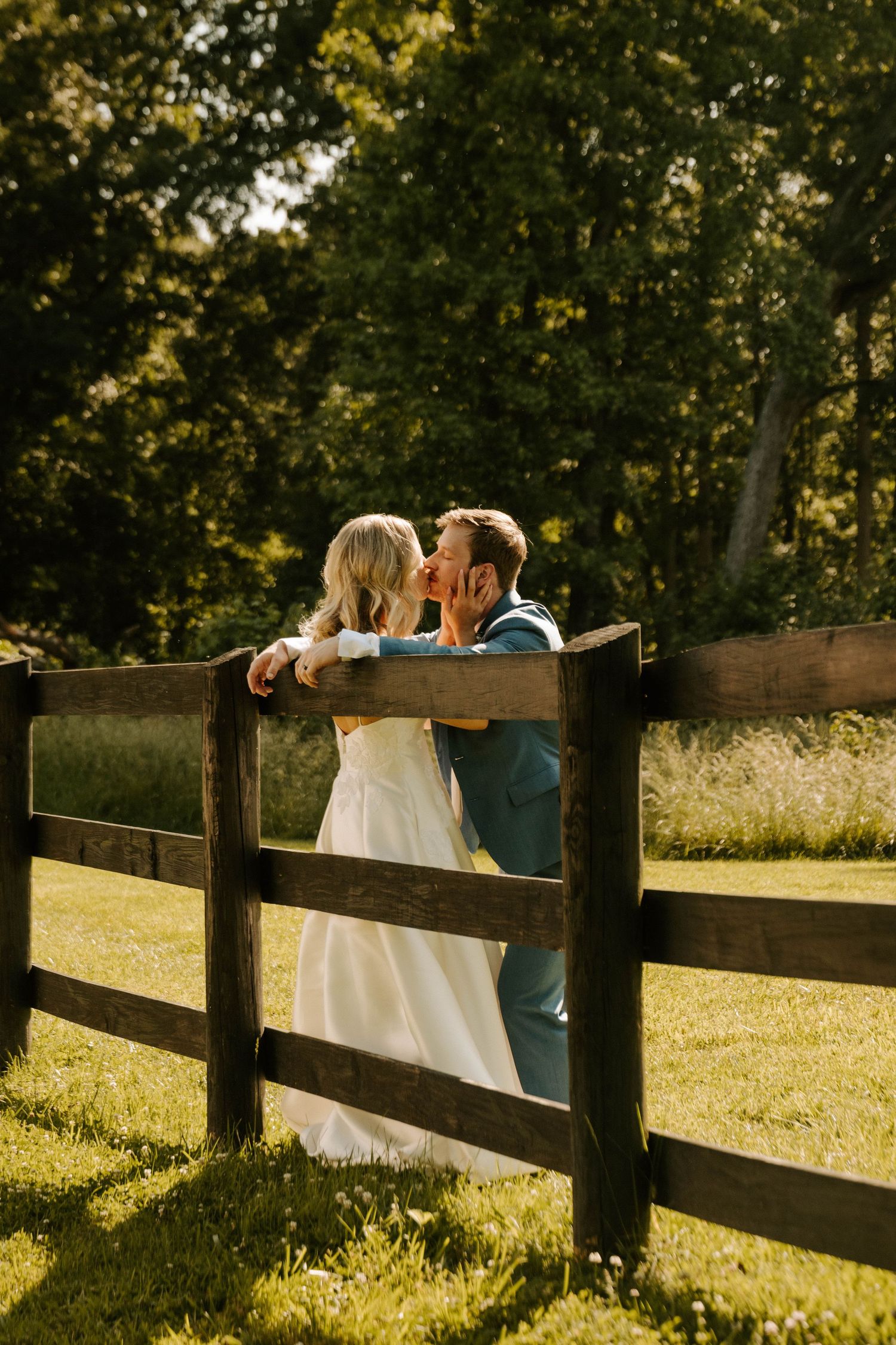 Couple sits on wooden fence together at sunset in golden summer field with trees in background.