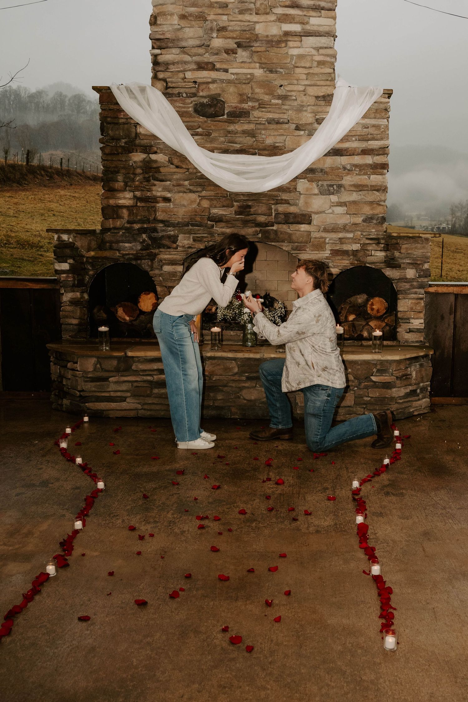 A romantic proposal scene unfolds against a stone wall backdrop with scattered rose petals and flowing white fabric.