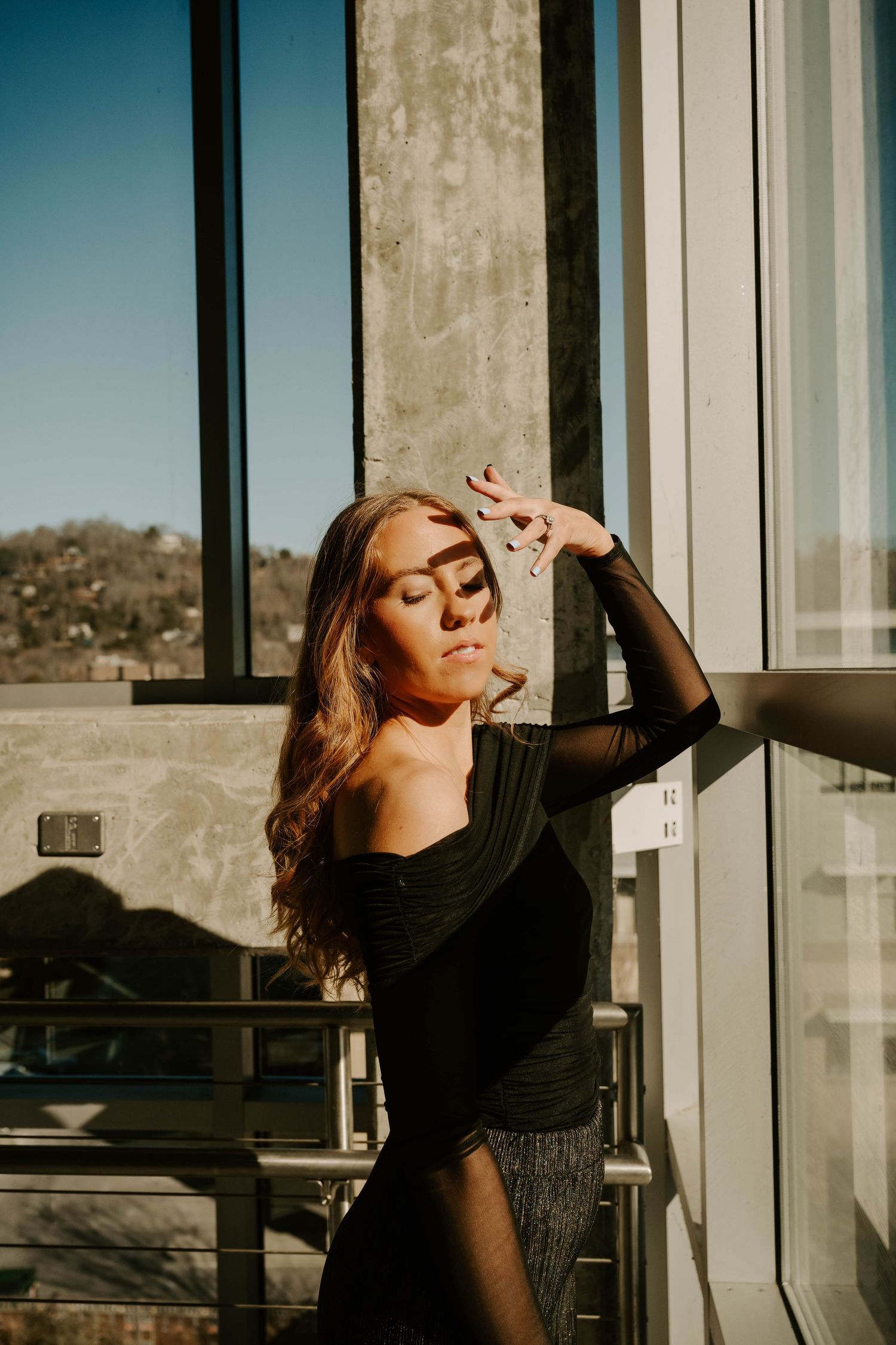 A woman in a black outfit poses dramatically by large windows in warm natural sunlight.