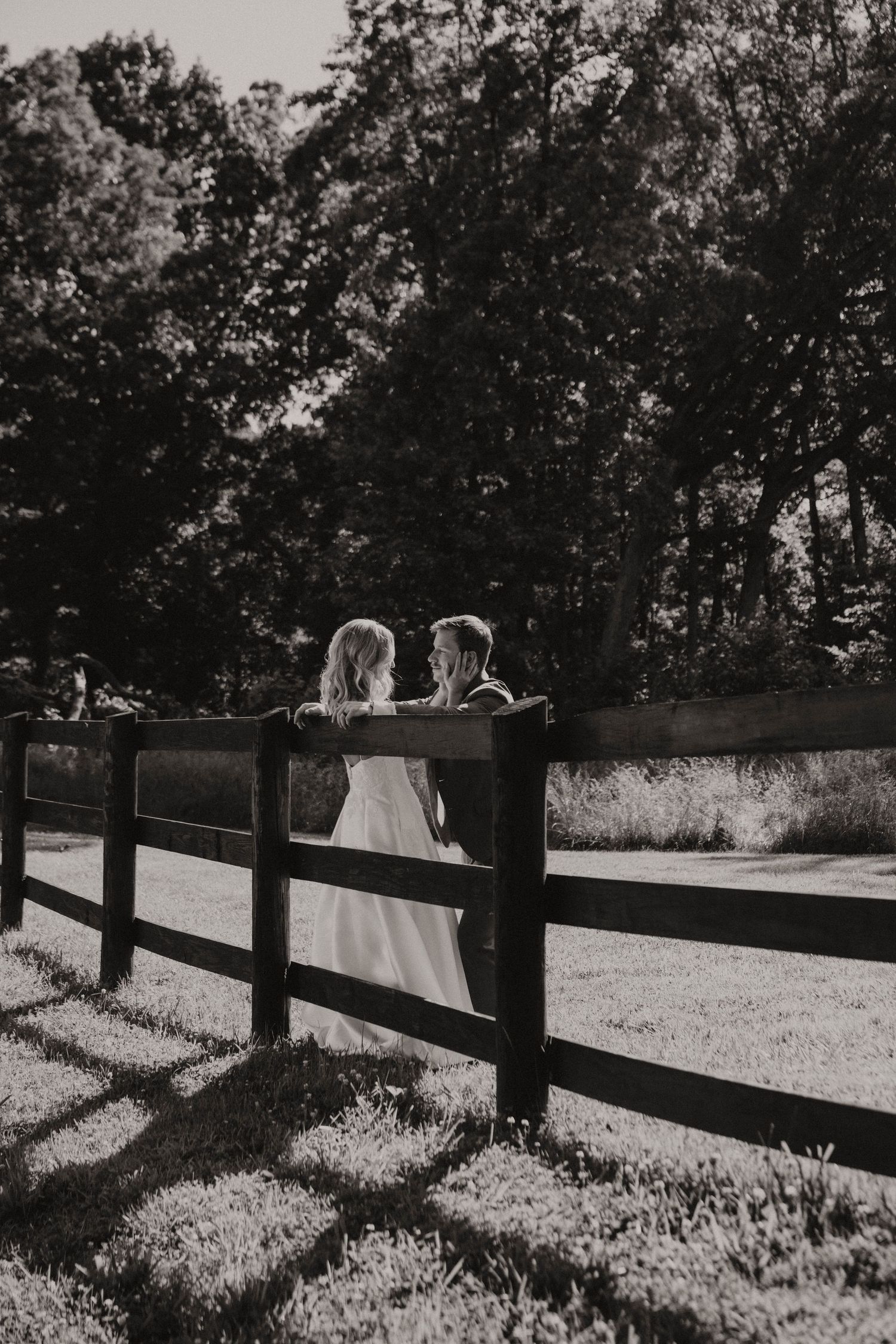 Silhouetted couple sits along wooden fence casting long shadows on sunlit grass in black and white rural scene.