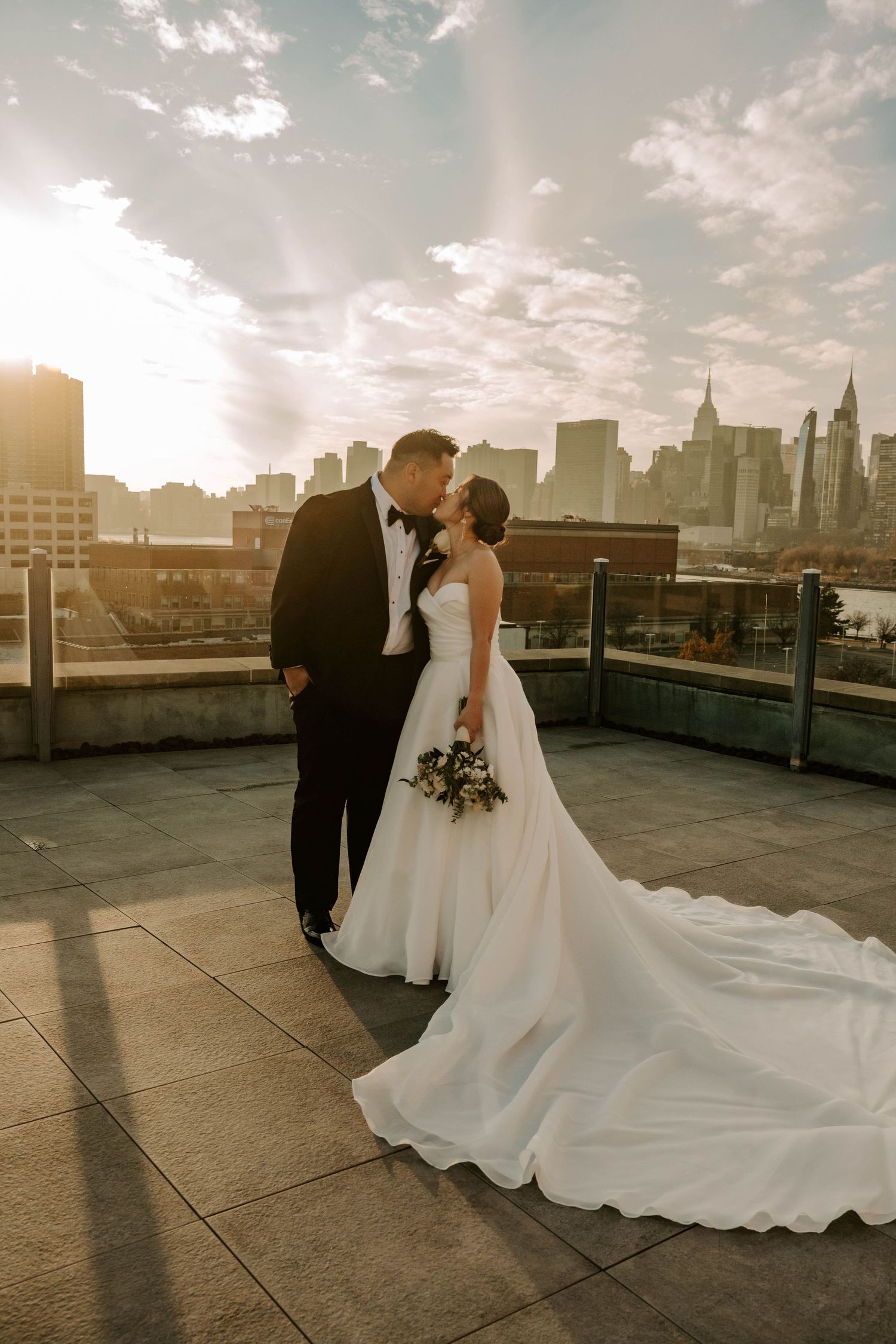 A romantic wedding couple shares a moment on a rooftop terrace with the New York City skyline at sunset.