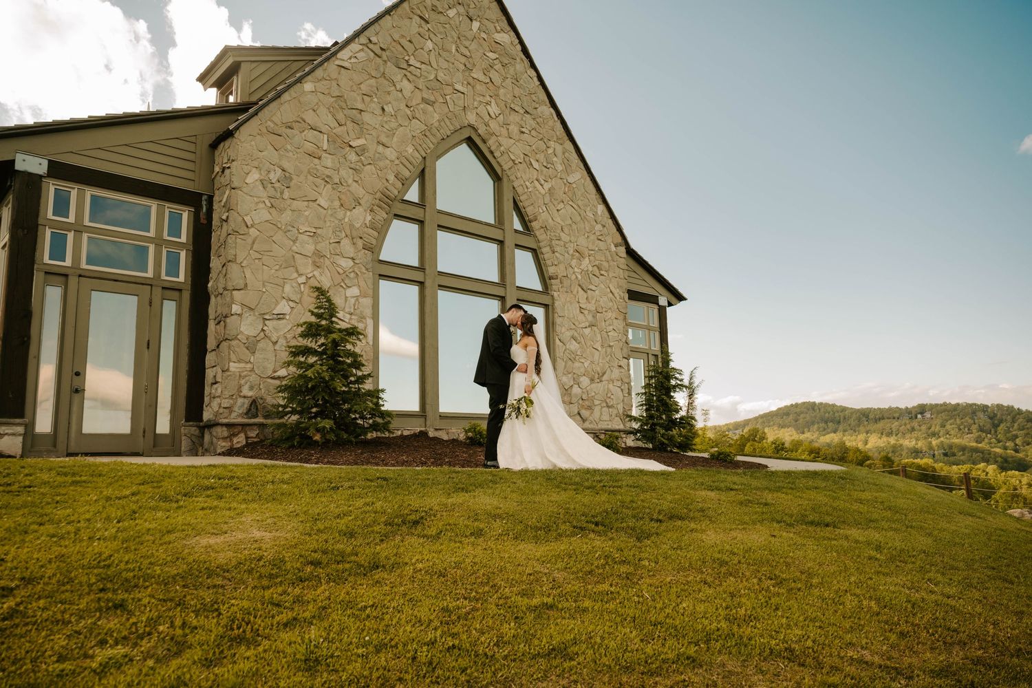 A couple stands outside a stone church with large windows on a grassy hilltop during a bright sunny day.