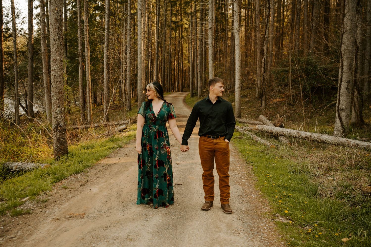A couple walks hand in hand on a dirt path through a forest of tall pine trees during golden hour.