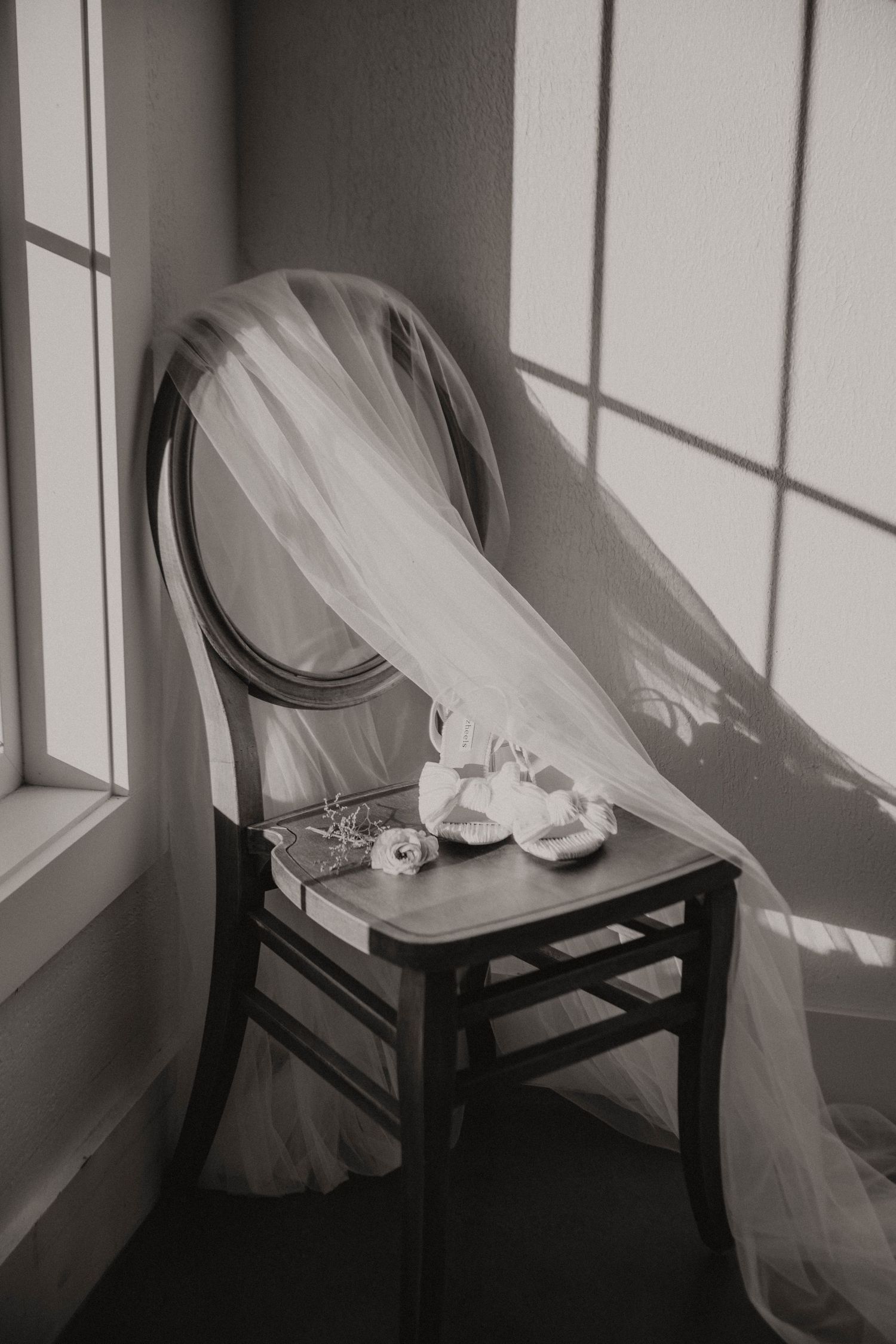 A romantic black and white photograph of a bridal veil draped over a vintage wooden chair near a window.