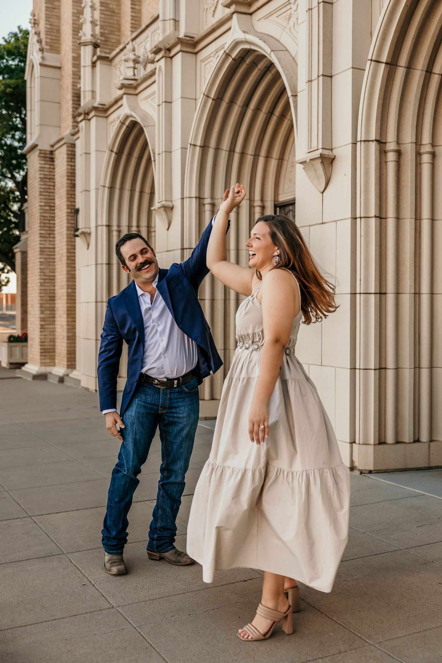 A couple in formal attire shares a playful moment under arched stone columns at an elegant venue.