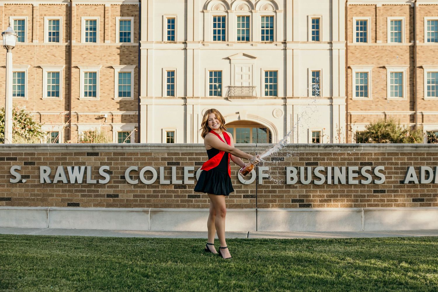 A figure in graduation attire poses in front of Rawls College of Business Administration building during golden hour.