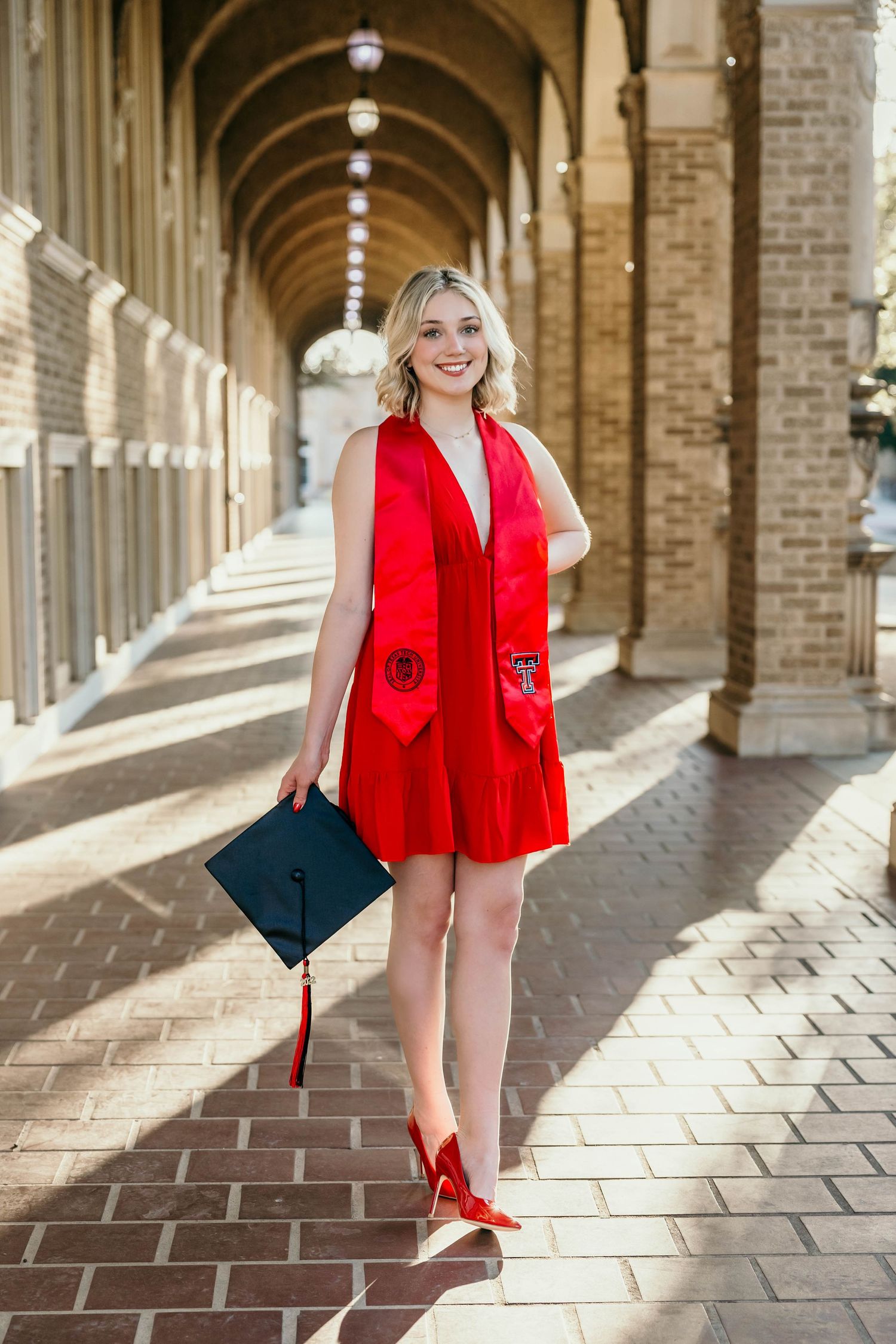 A red sleeveless romper outfit with matching red shoes and black handbag photographed under an arched colonnade walkway.