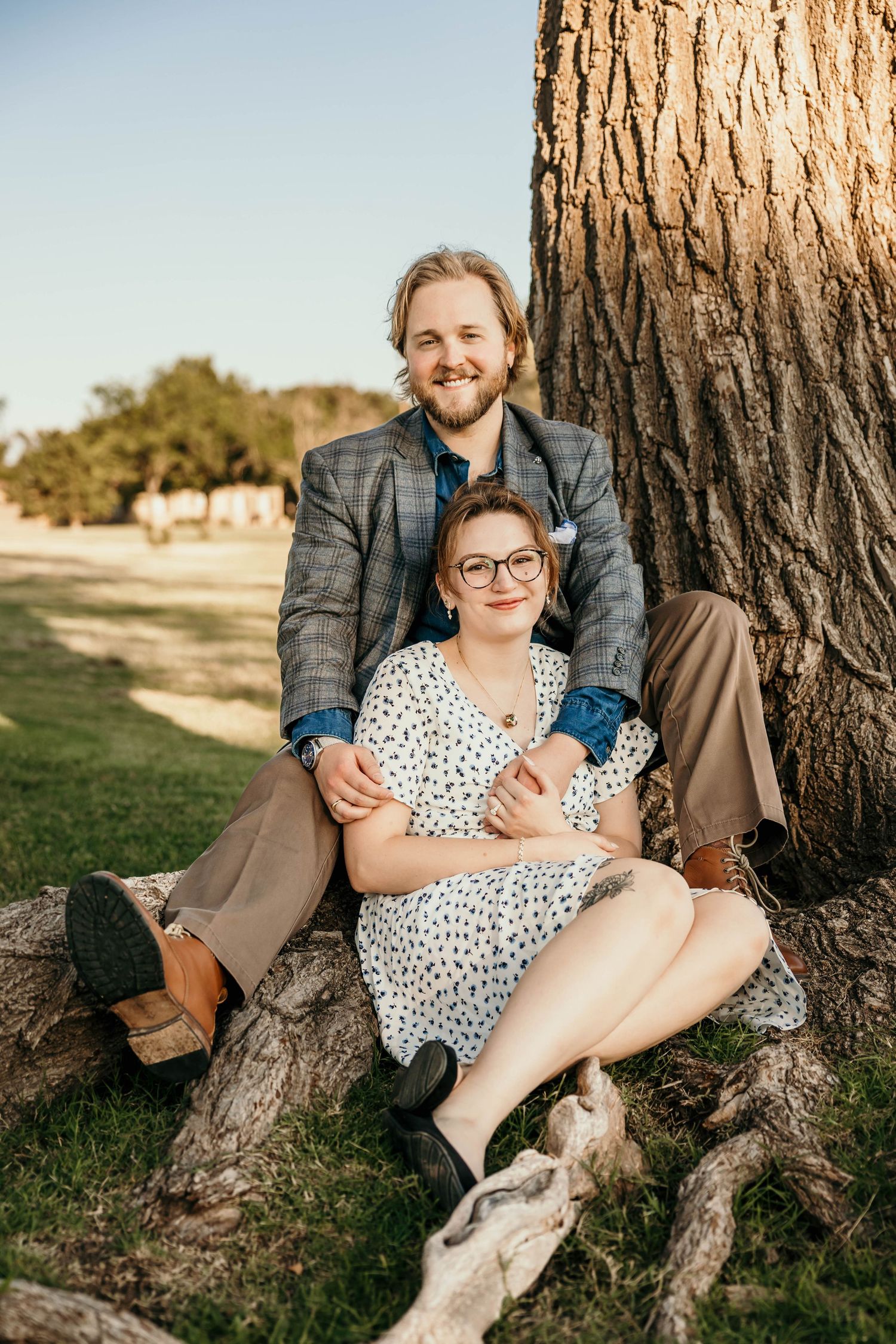 A couple sits together against a large tree trunk in a park during golden hour wearing casual formal attire.