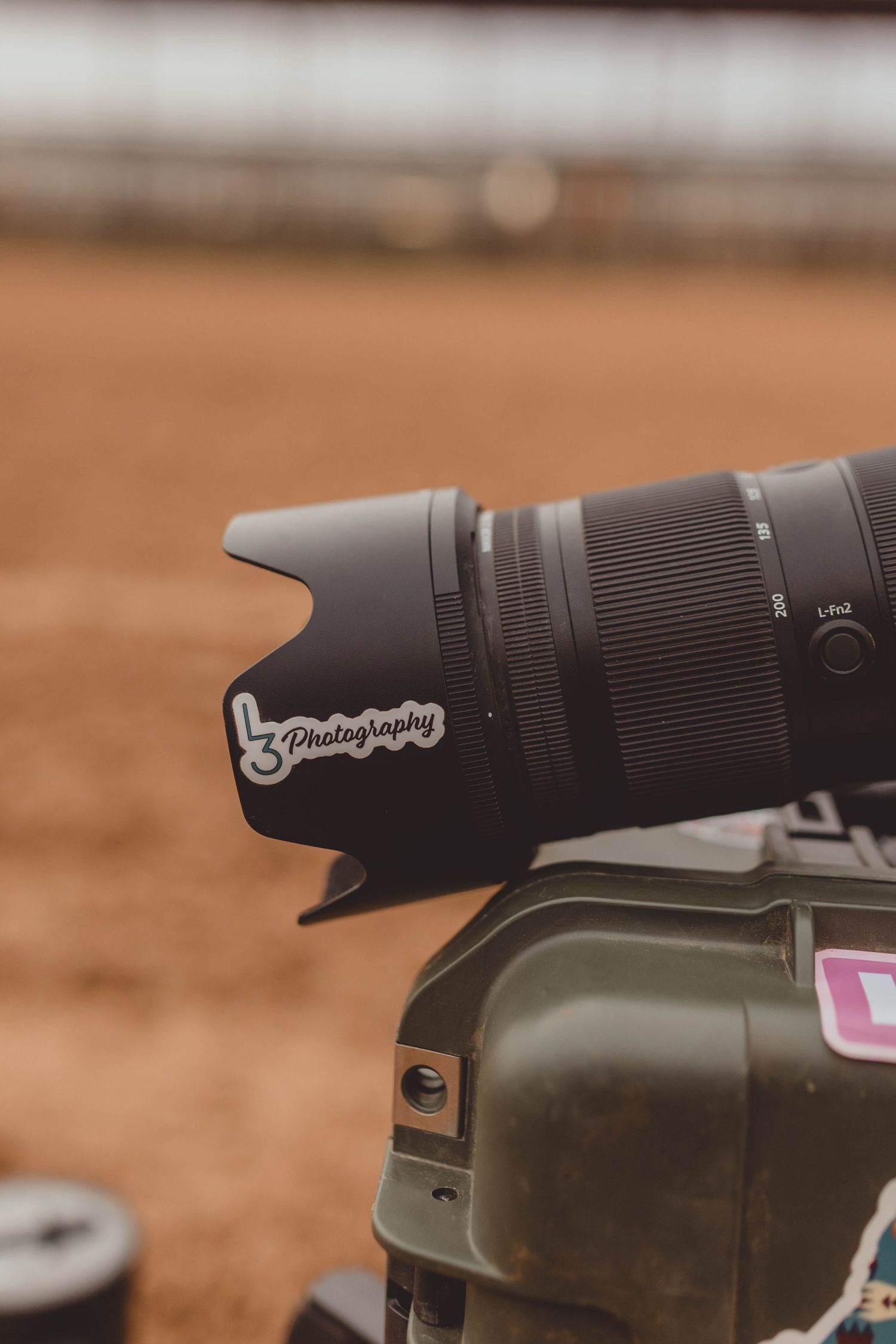 Close-up of a black camera telephoto lens with an L3 Photography sticker, mounted outdoors with a blurred dirt background.
