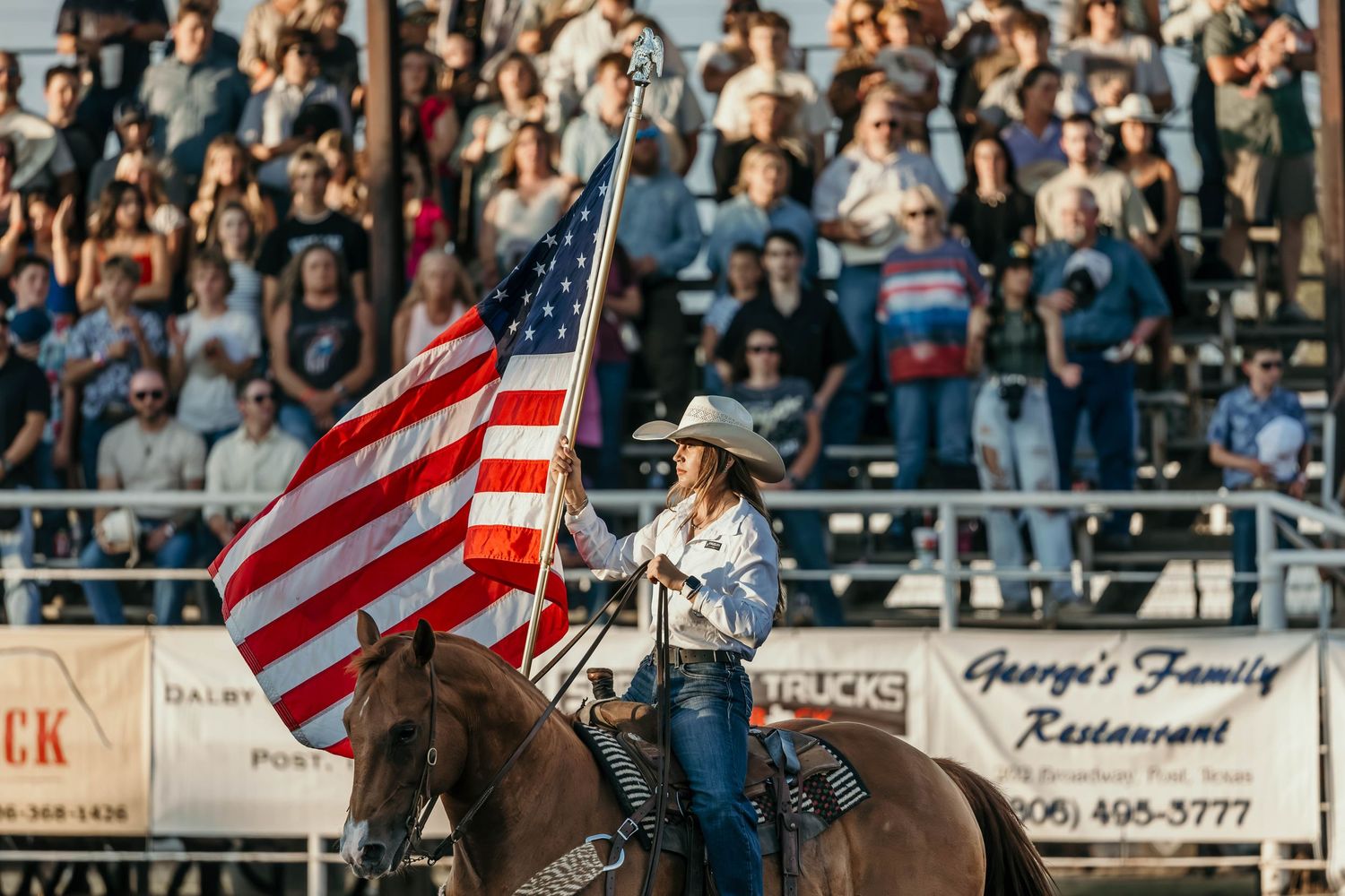 A cowgirl on horseback carries a large American flag at a rodeo arena filled with spectators.