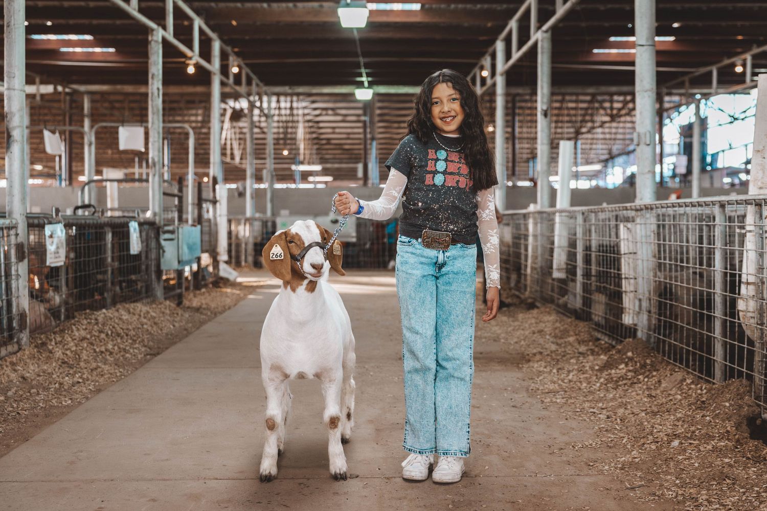 Inside a barn aisle lit by overhead lights showing animal pens with straw bedding and wire fencing.