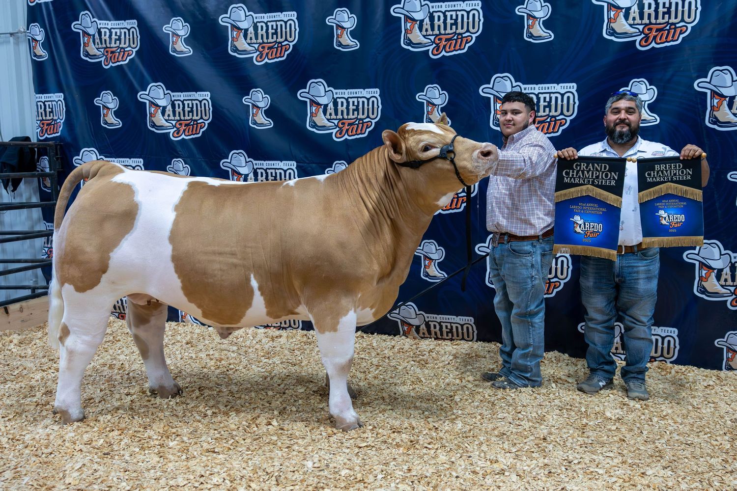 A brown and white bull is shown at a livestock show with handlers posing against a backdrop with repeating logos.