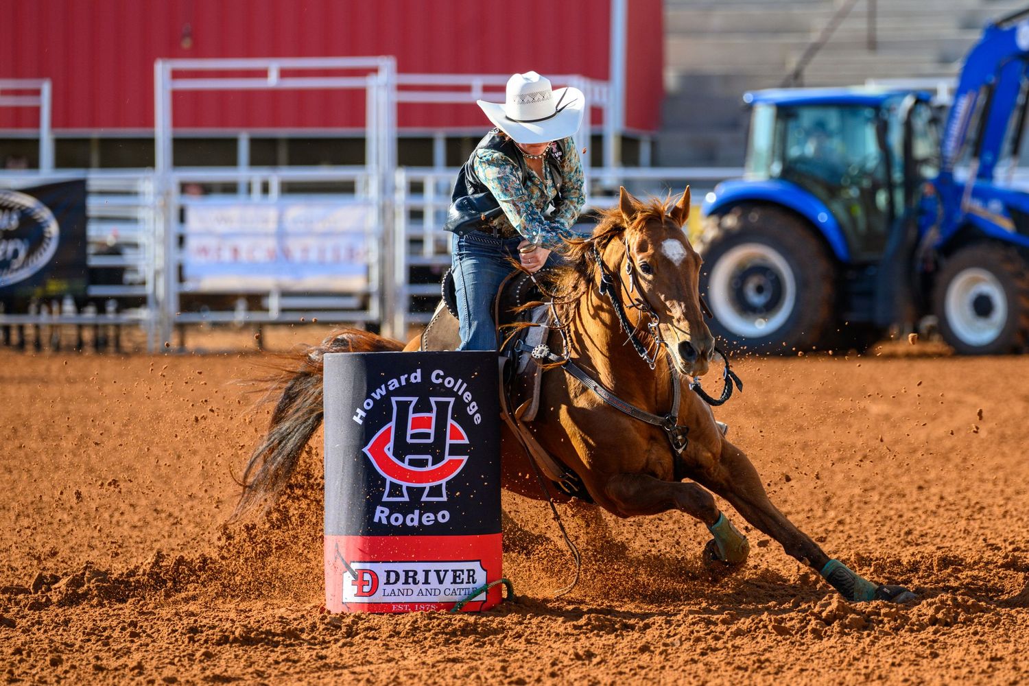 Barrel racer on chestnut horse competing at Howard College Rodeo, kicking up dirt around a turn barrel indoors.