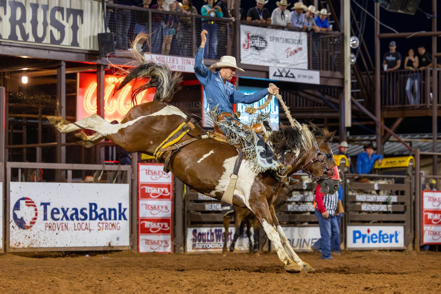 Cowboy rides bucking bronco at Texas Bank rodeo event under bright arena lights.