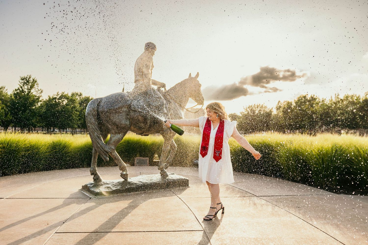 A person in white dress dances next to a bronze equestrian statue at sunset with sun rays shining through trees.