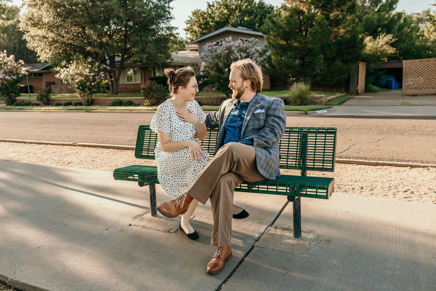 Two people sitting and talking on a green park bench on a sunny day with trees in the background.