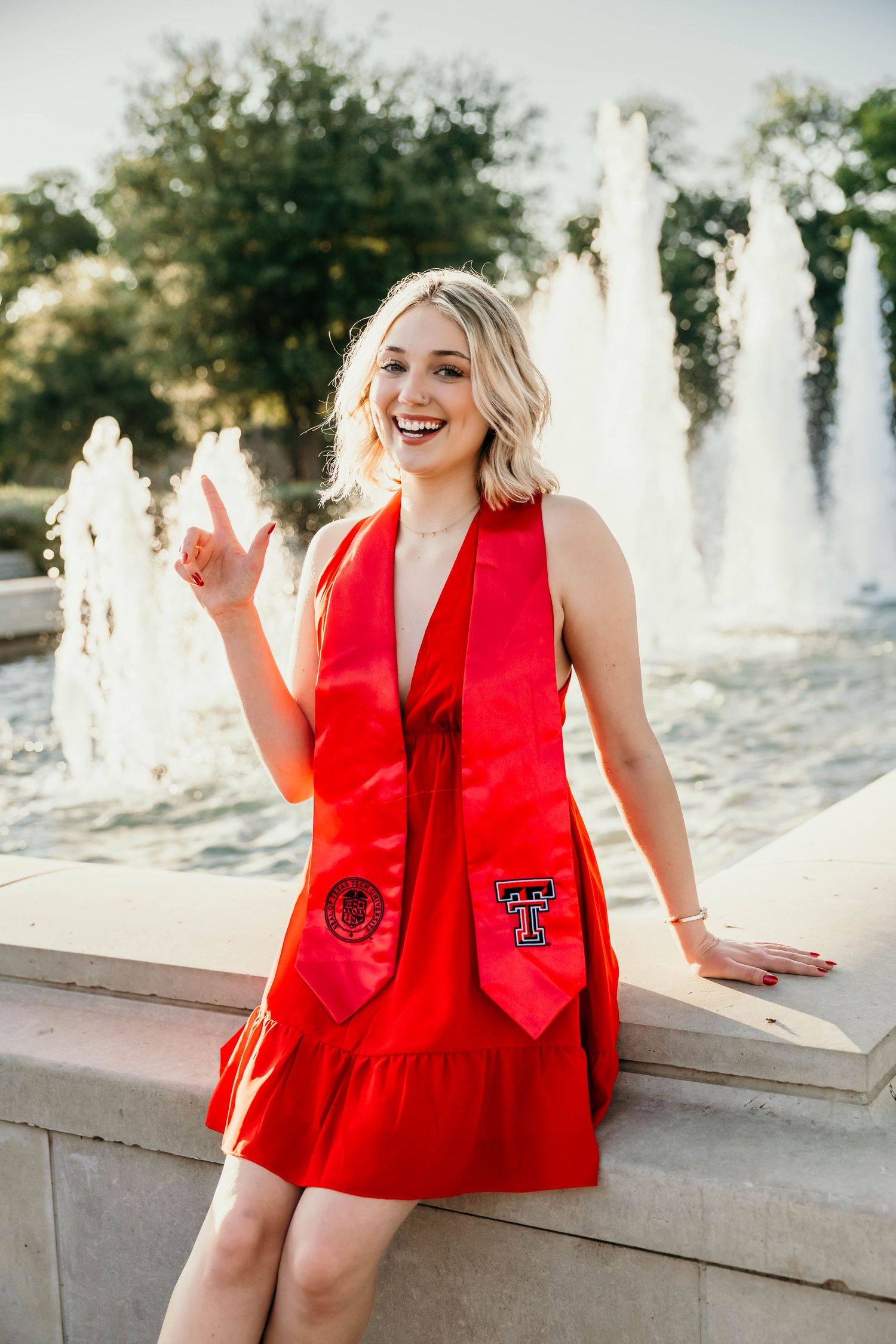 Graduate in red dress poses joyfully by water fountain with Texas Tech stole during sunset graduation photoshoot.