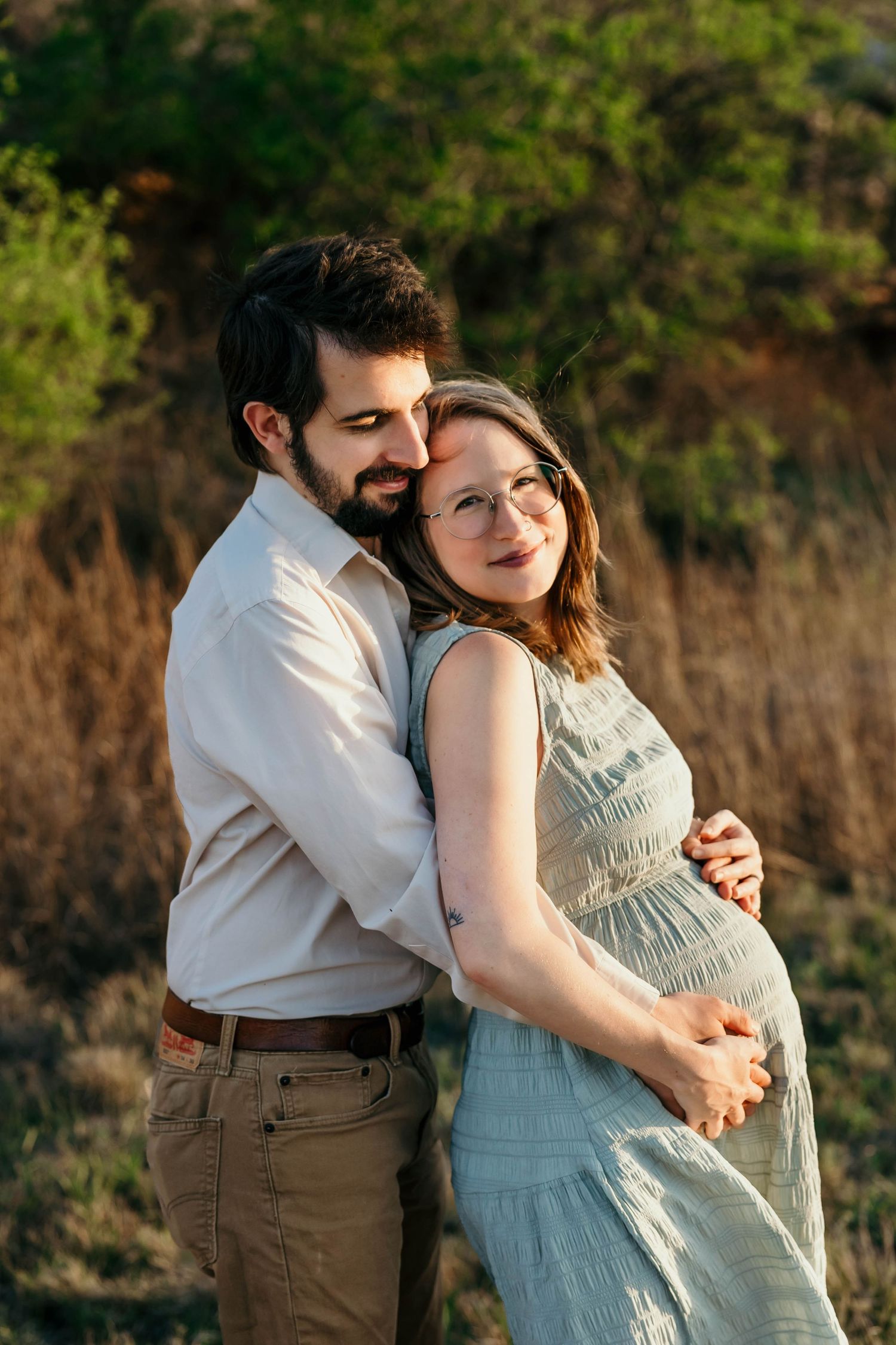 A couple embraces outdoors during sunset, with the woman wearing a light blue dress cradling her pregnant belly.