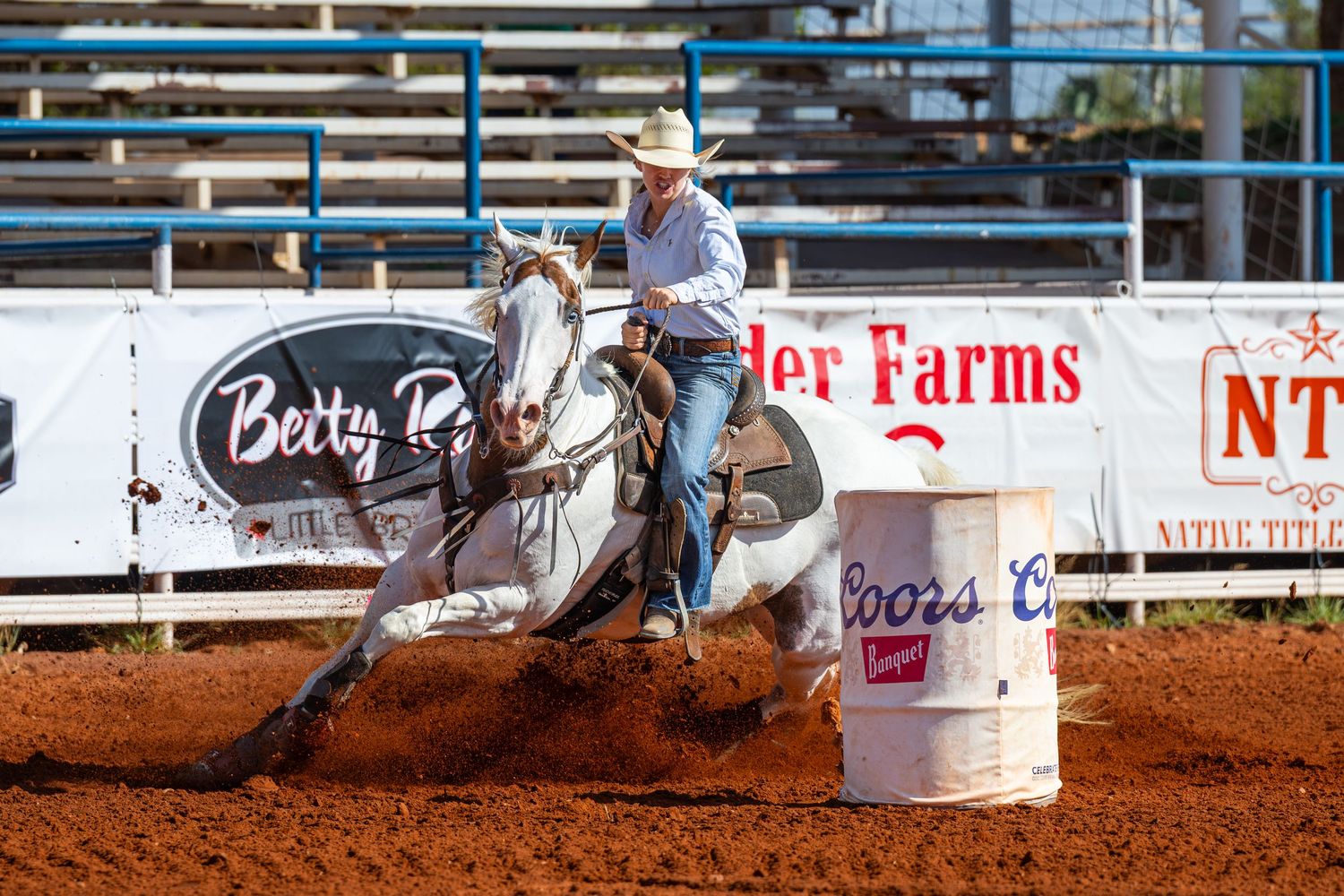 Barrel racer on white horse navigating turn at rodeo arena with Coors Light barrel and Betty R Farms signage.