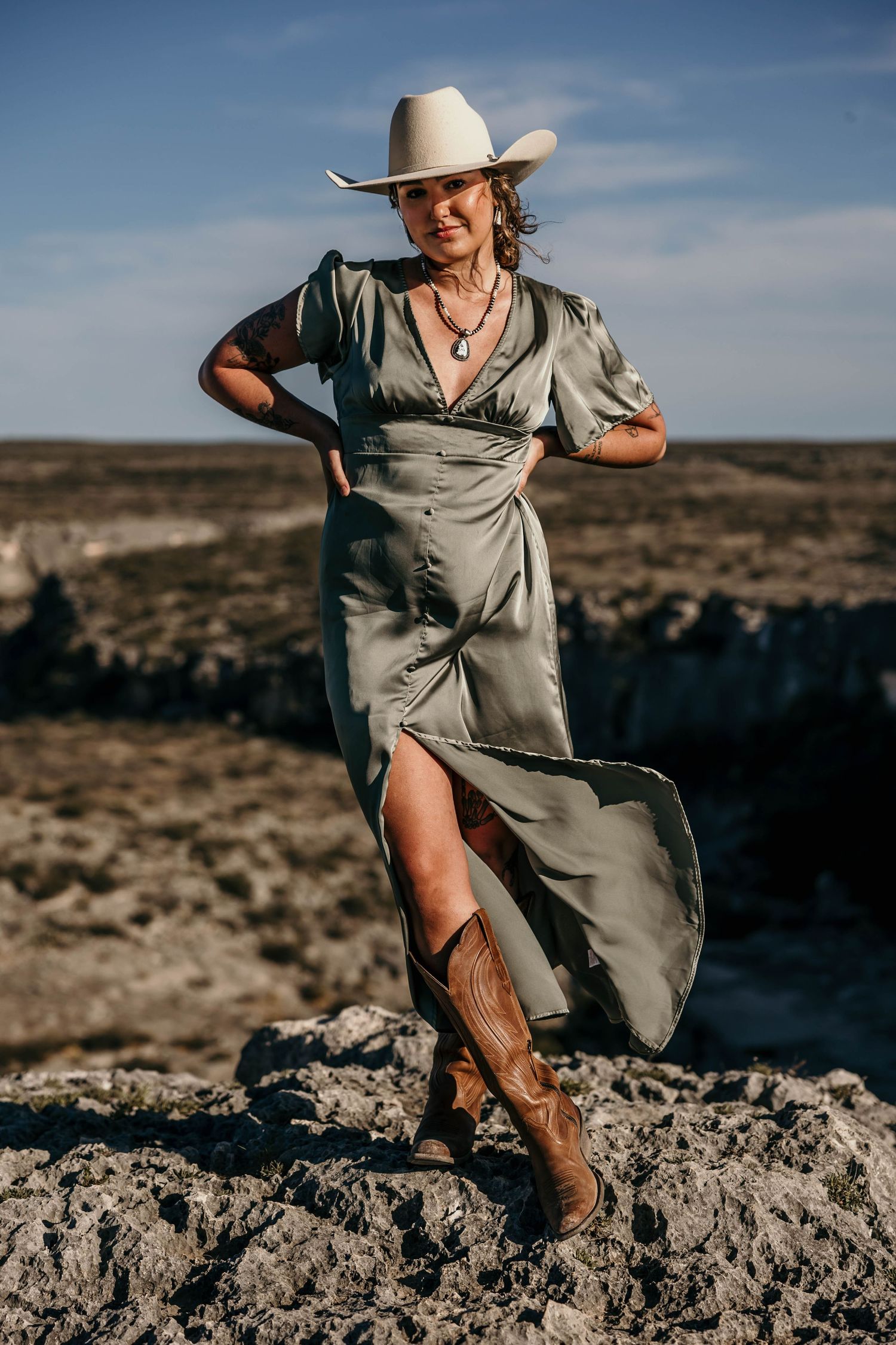 A person in a flowing sage dress and cowboy hat poses dramatically on rocky desert terrain against a blue sky.