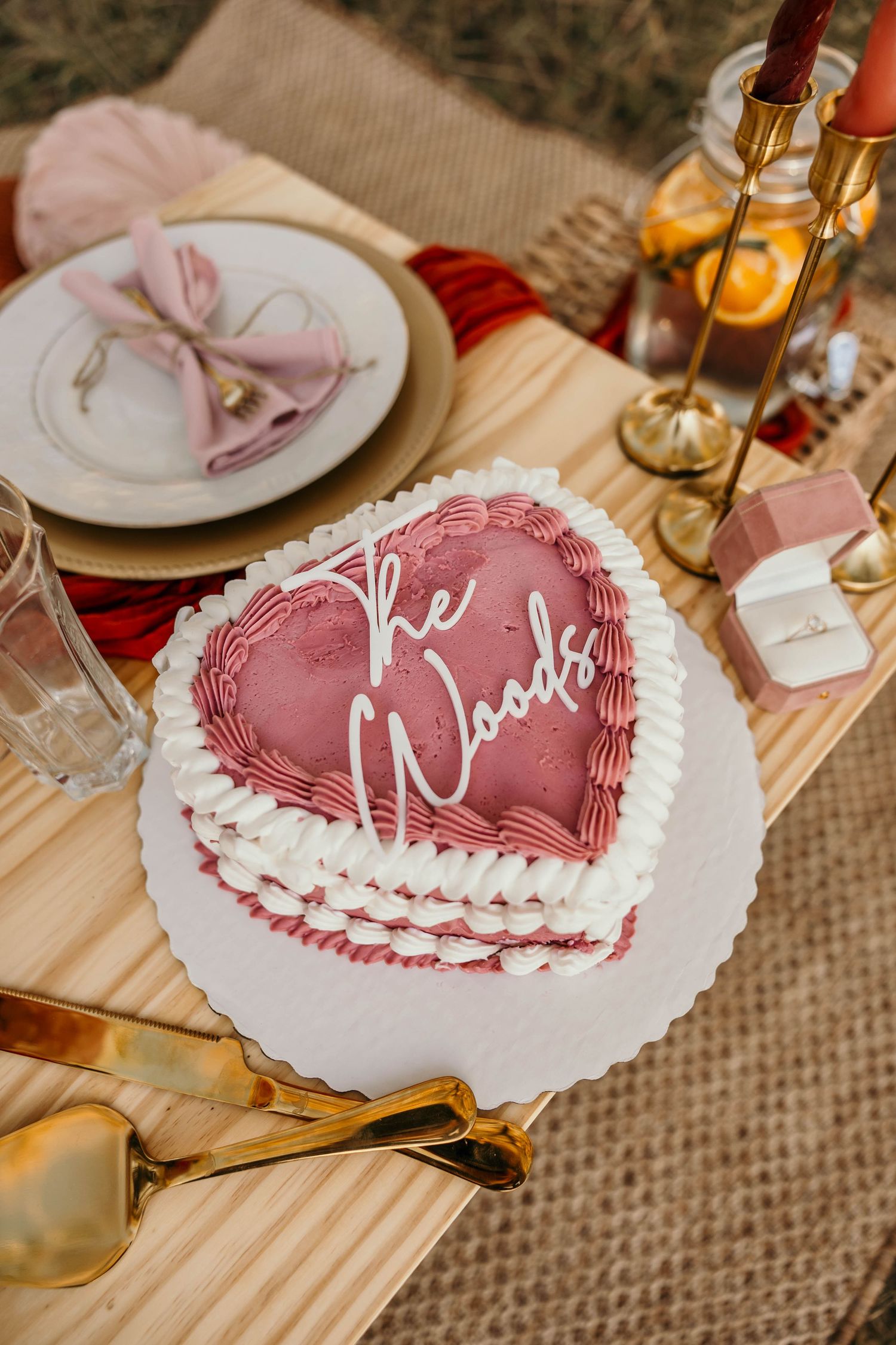 Heart-shaped pink and white decorated Valentine's cake displayed on rustic table settings with candlelight and burlap details.