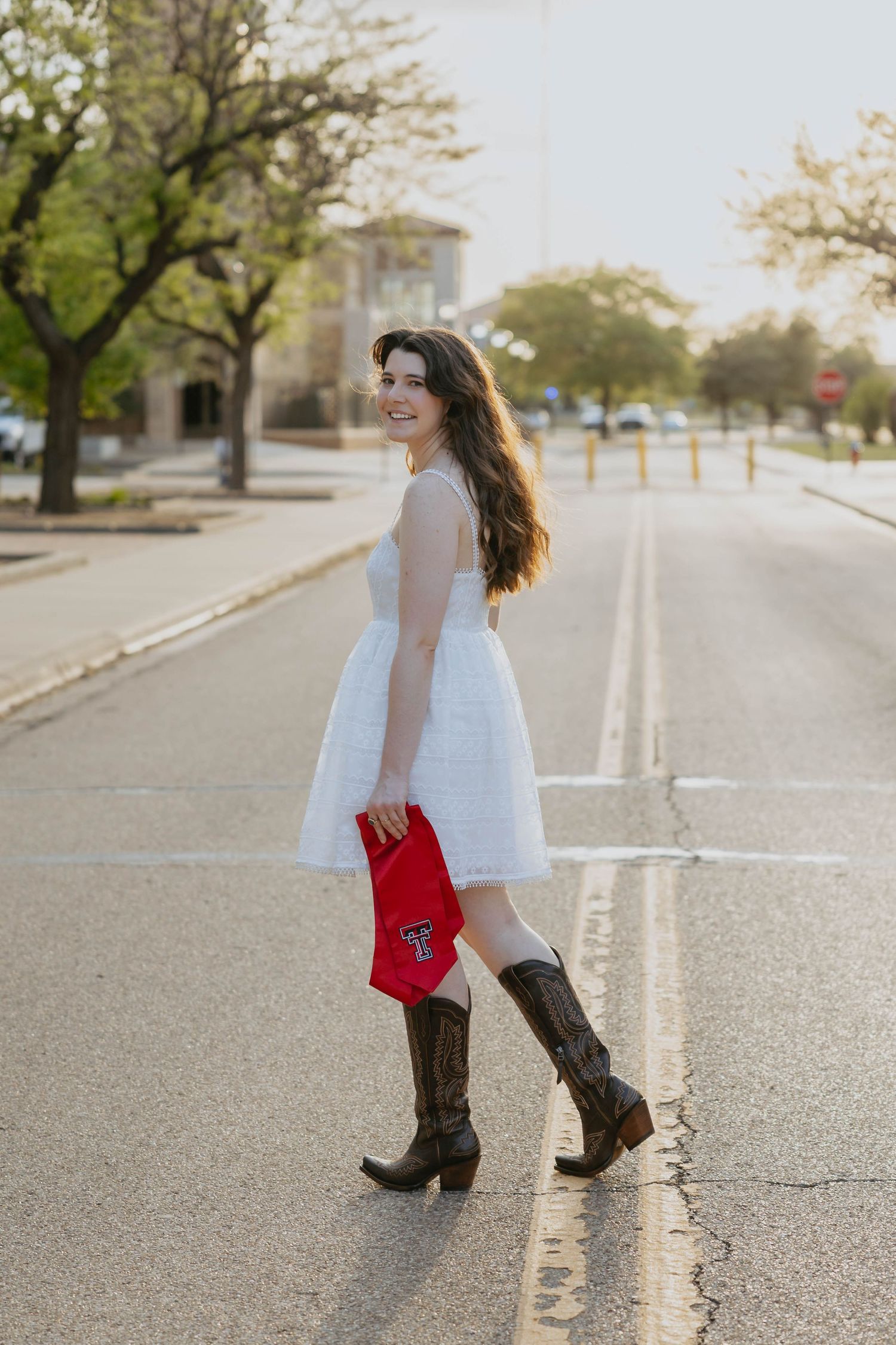 Someone in a white dress and brown cowboy boots poses on a sunny street with a red bag.