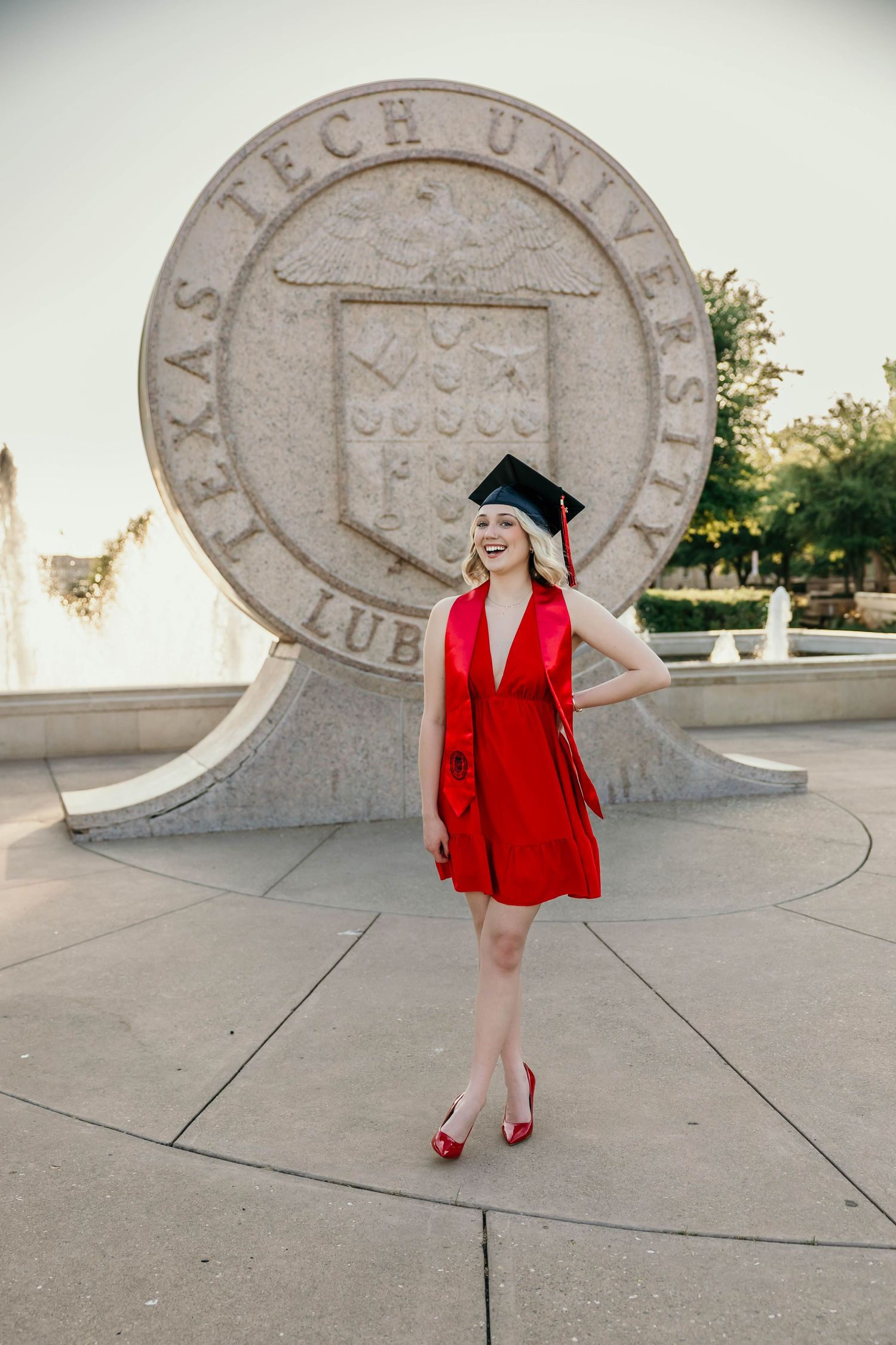 Graduate poses near campus seal in red blazer and graduation cap during outdoor photo session at Texas Tech University.