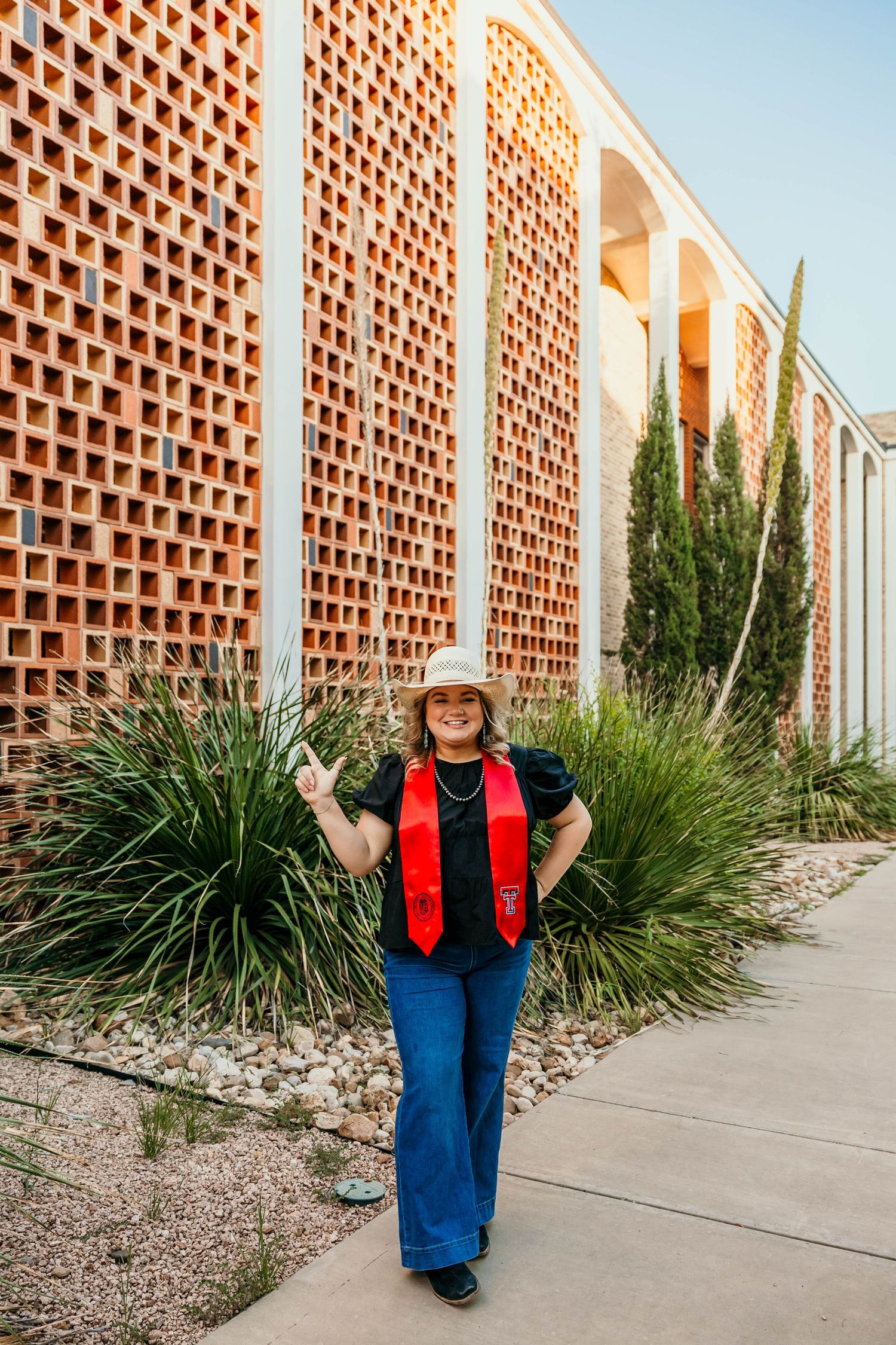 Person in red vest and denim poses by modern brick wall with decorative lattice pattern and desert landscaping.