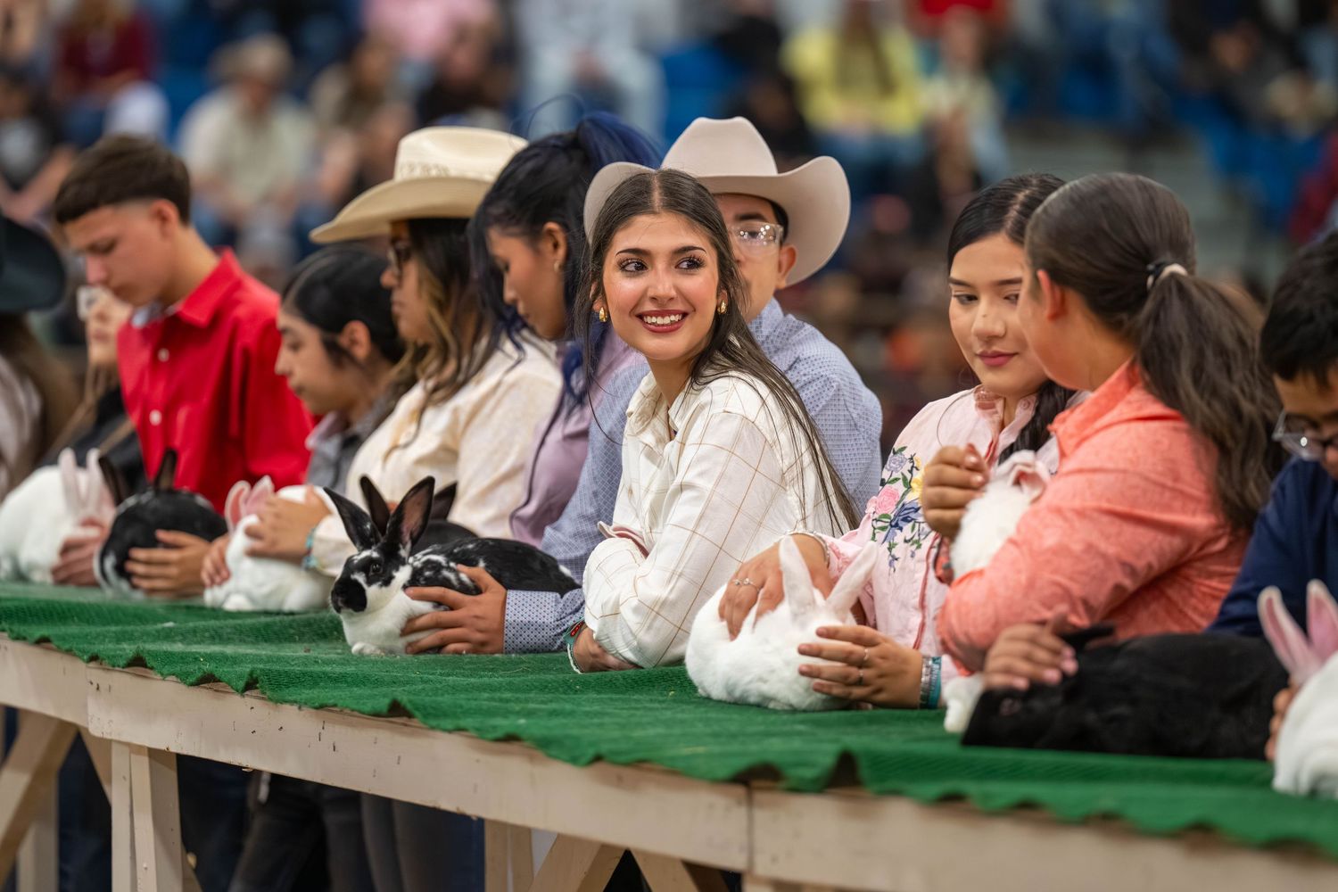 Young participants hold rabbits at a county fair rabbit show, wearing cowboy hats and casual attire indoors.