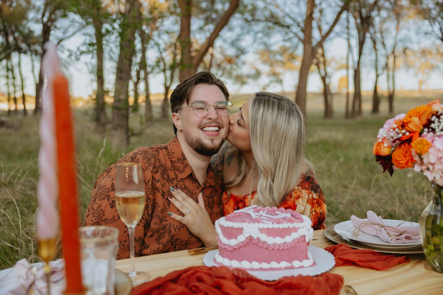 A romantic outdoor picnic setting at sunset with pink and orange decor, flowers and cake on a rustic wooden table.