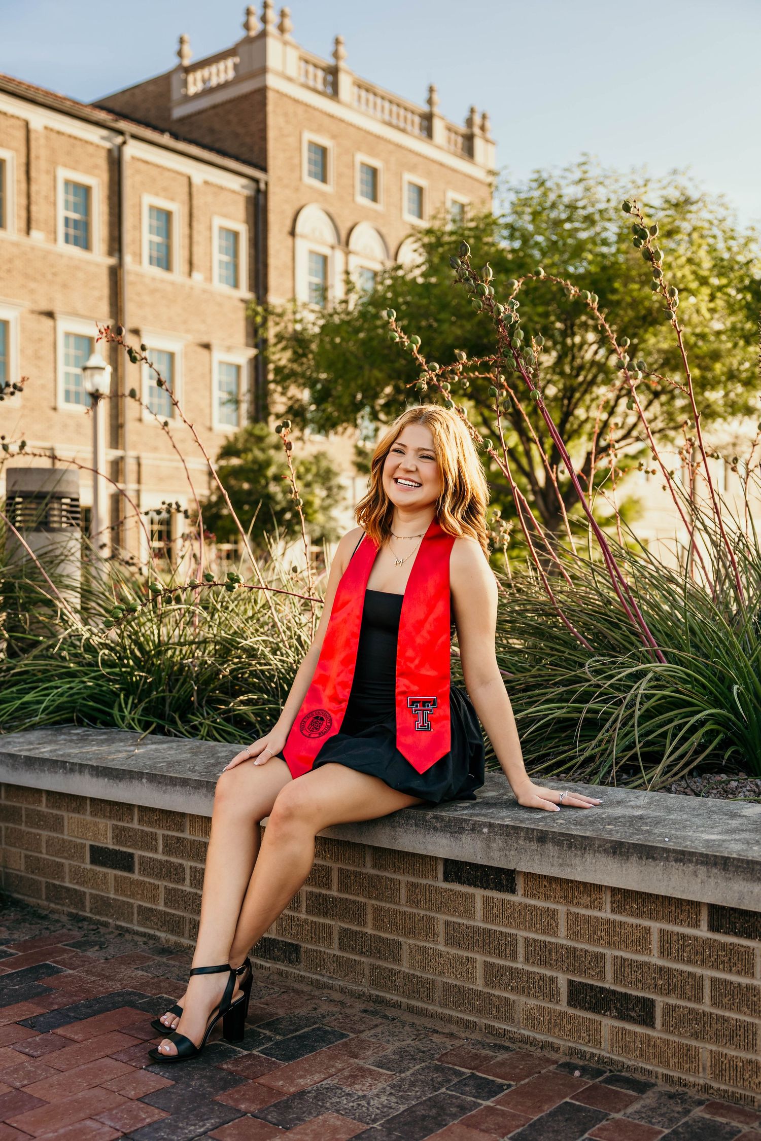 Person in red and black outfit sitting on brick wall with buildings and trees in background during golden hour.