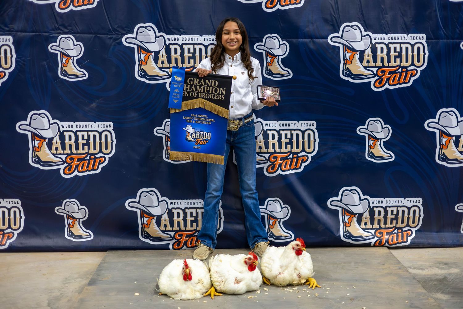 Winner poses with championship banner and trophies at Laredo event with large logo backdrop and cotton bales.