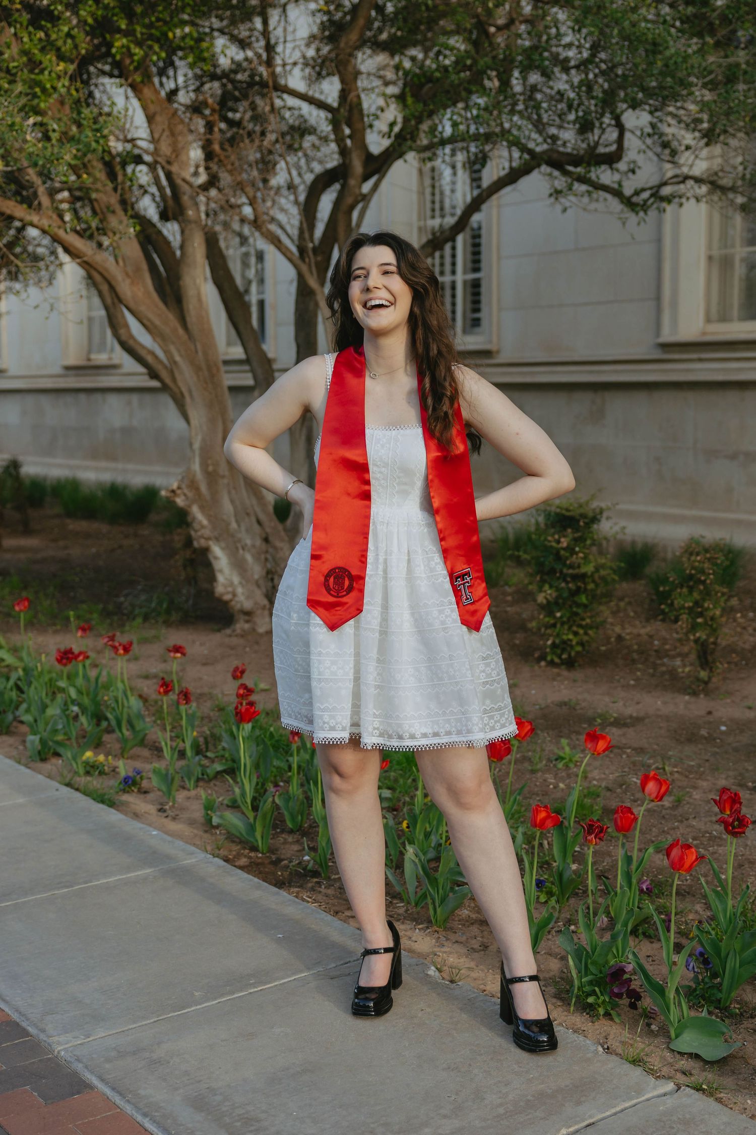 A person wearing a white dress and red vest poses playfully in a garden with blooming red tulips and trees.