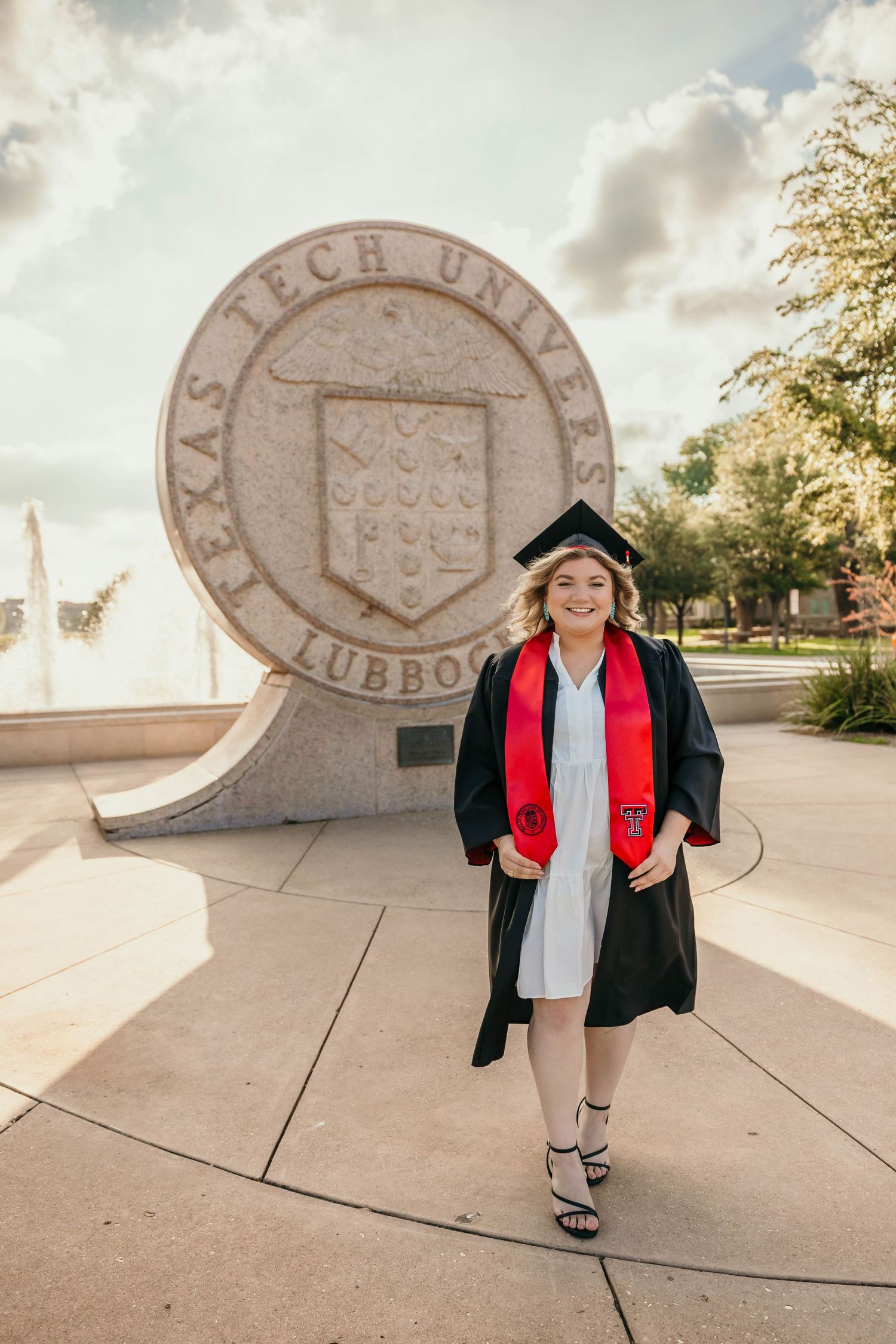Graduate in cap and gown poses in front of Texas Tech University seal on campus with fountain in background.