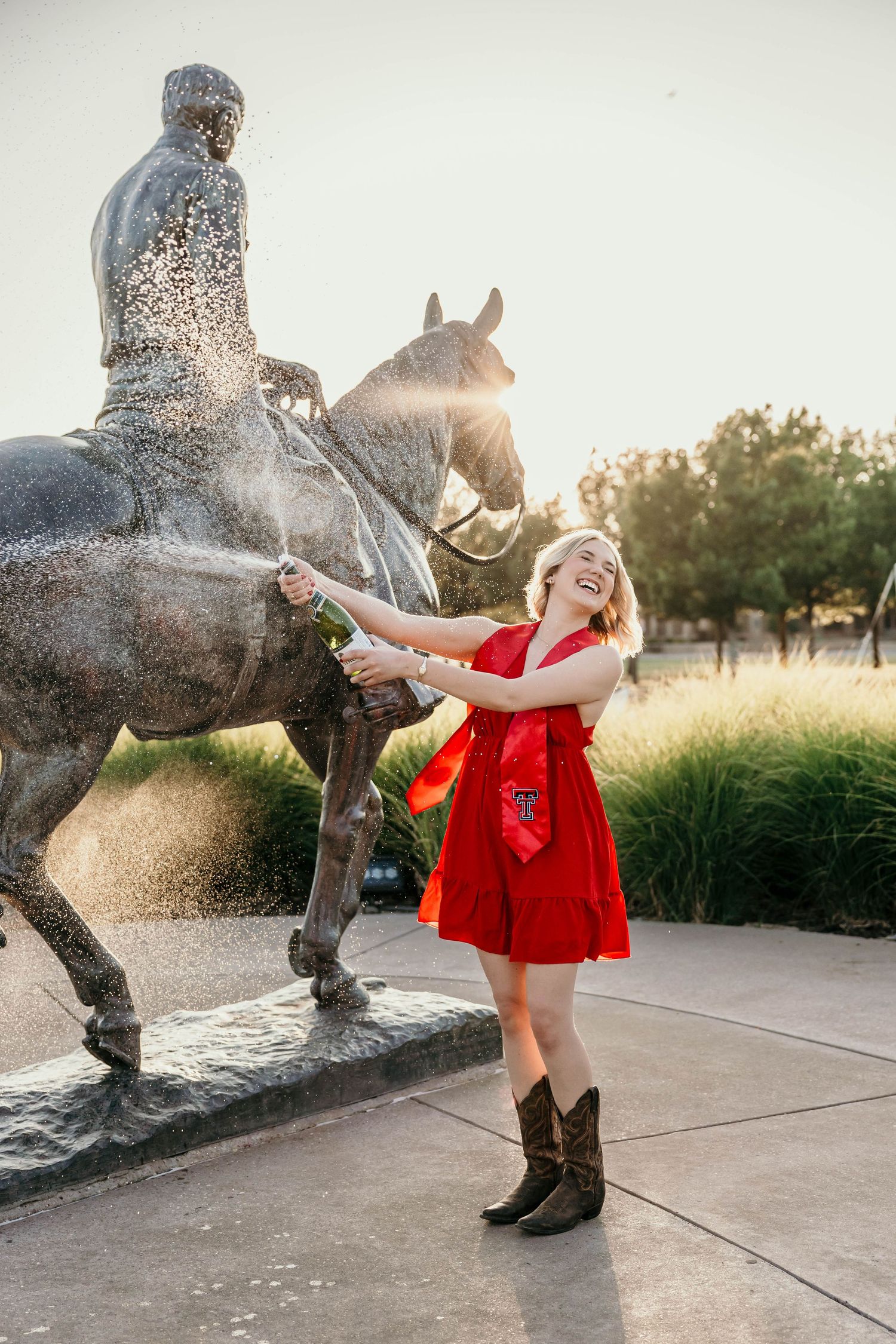 Graduate in red dress and cowboy boots poses playfully with bronze horse statue during outdoor photoshoot.
