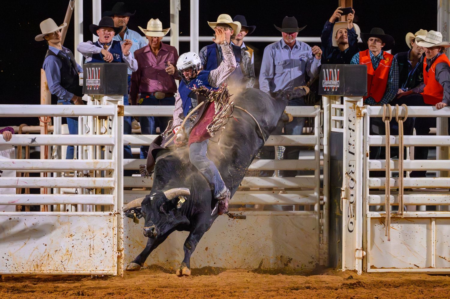 A cowboy rides a bucking bull at a rodeo arena at night, with spectators watching behind metal fencing.
