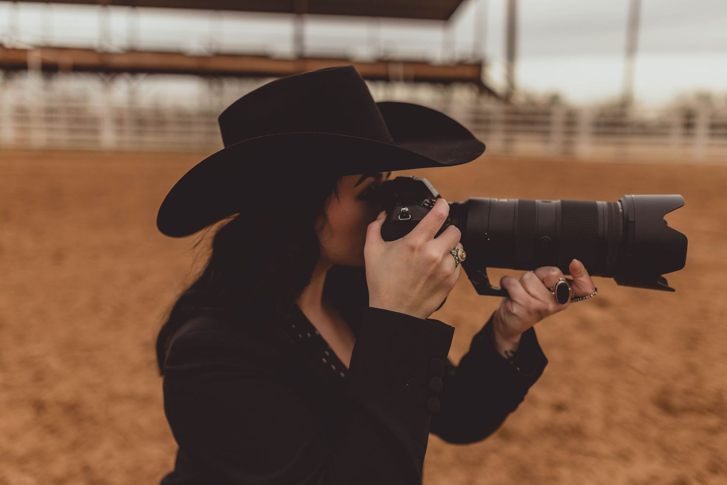 Woman in black cowboy hat photographing with DSLR camera at outdoor rodeo arena.