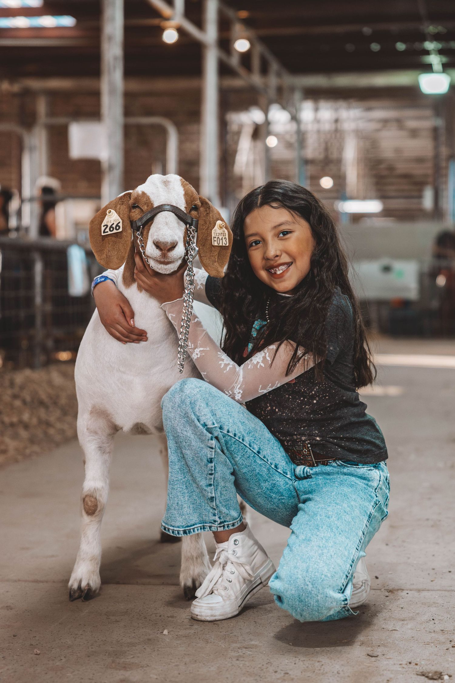 A friendly white and brown goat with a collar poses alongside someone wearing jeans in a rustic indoor setting.