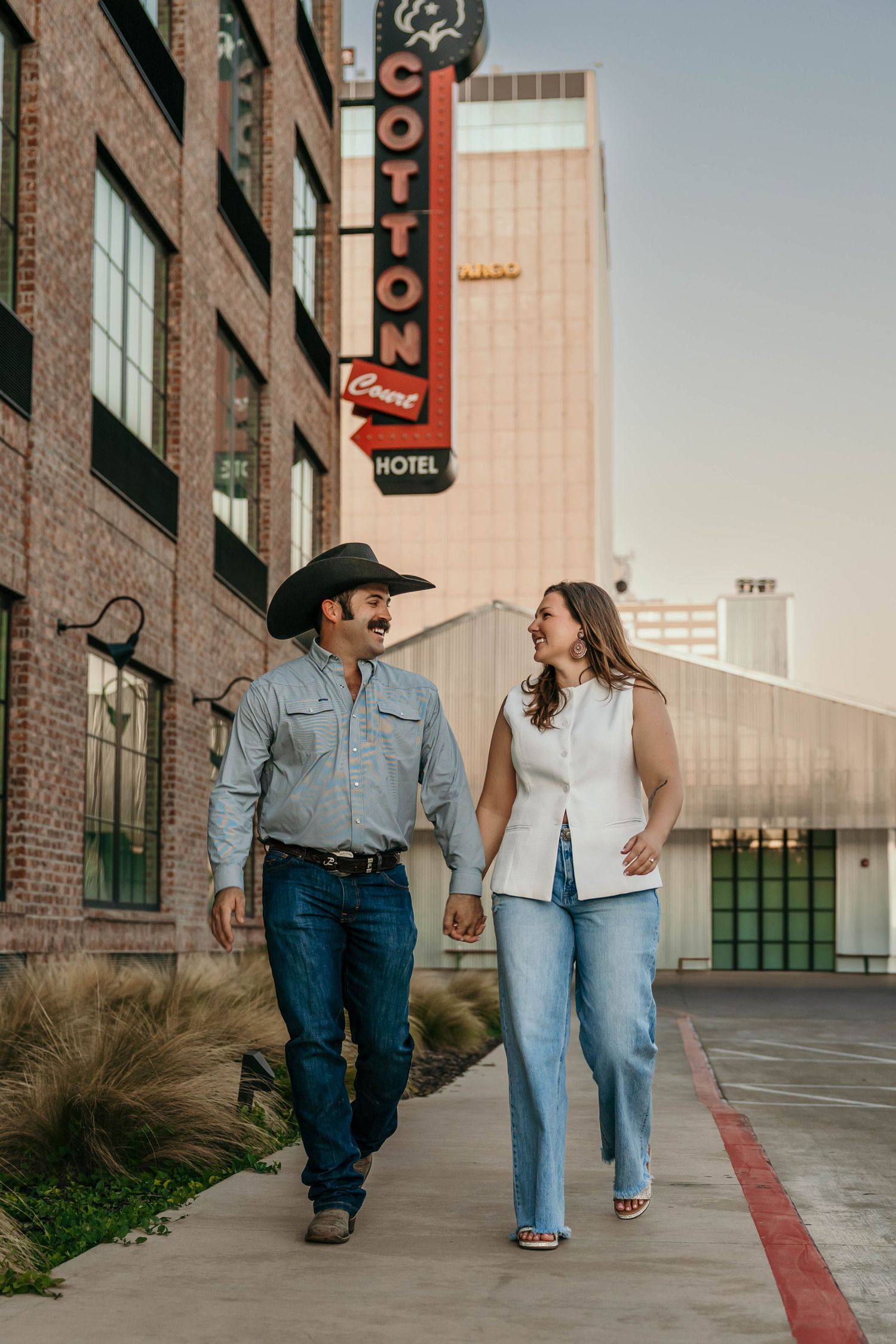 A couple in denim walks together down a city sidewalk with brick buildings and vintage hotel signs in the background.