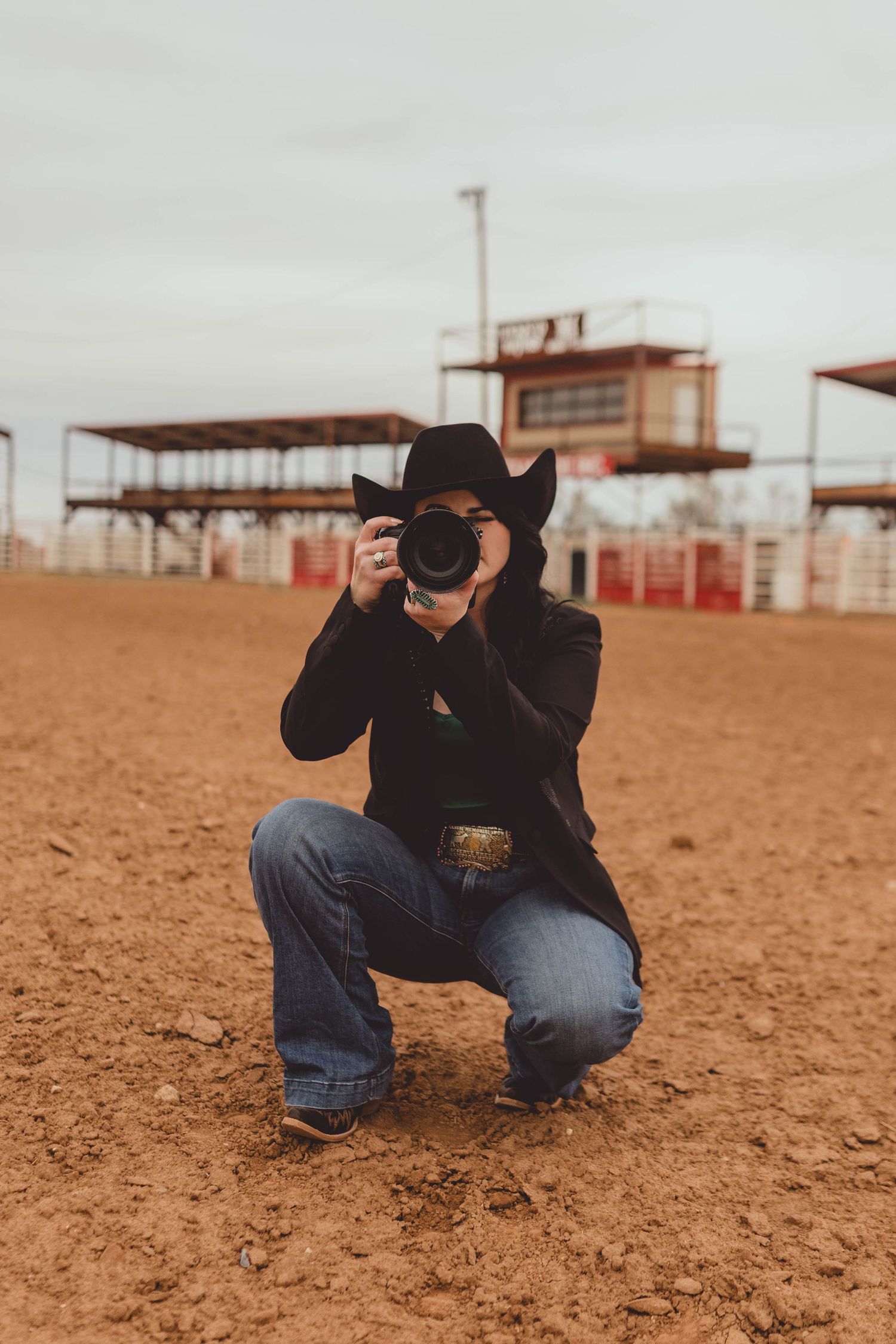 Photographer in black crouches low to the ground with camera in dusty arena setting.