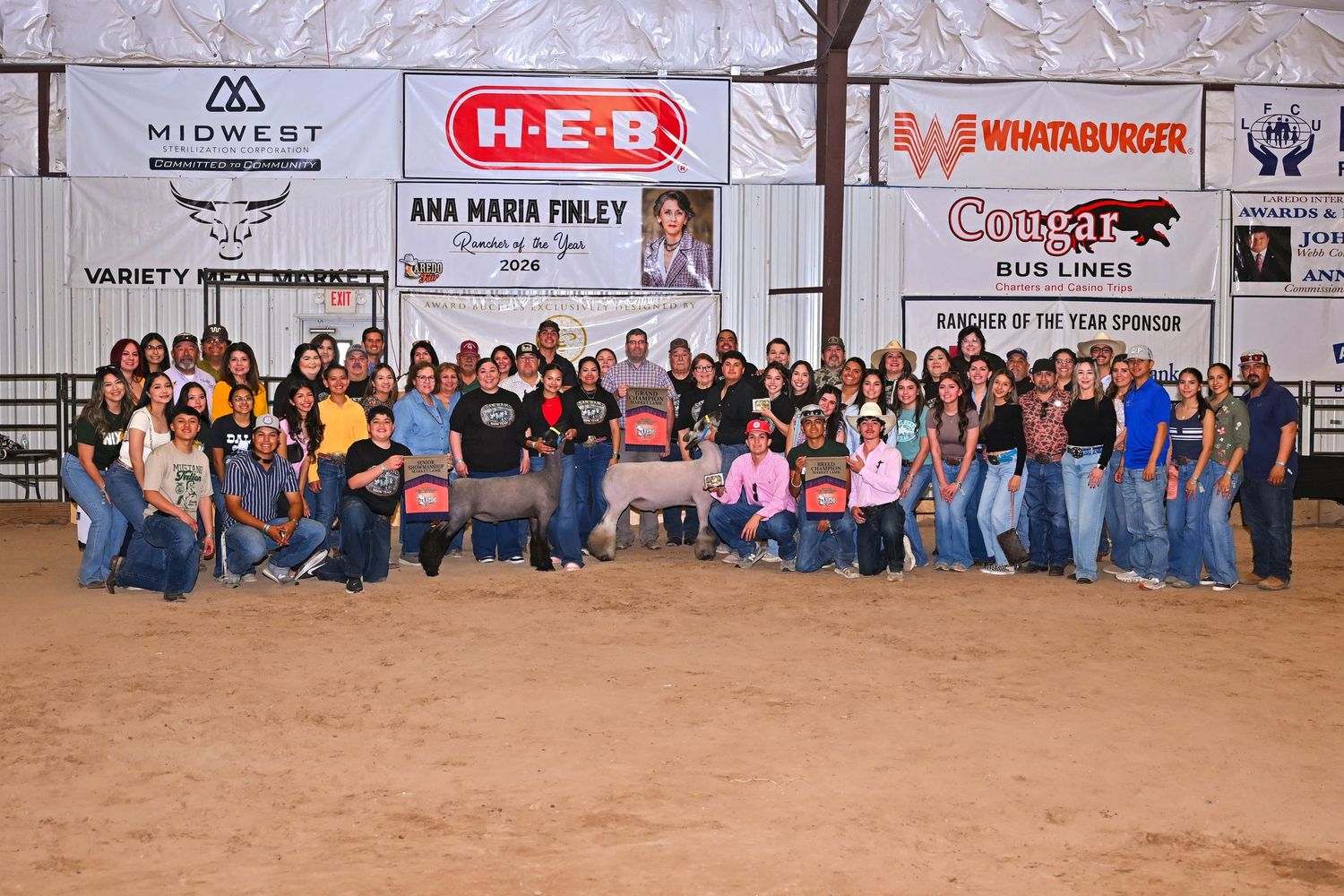 Large group photo at an indoor livestock show with a white goat, sponsored by H-E-B, Whataburger, and Cougar Bus Lines.