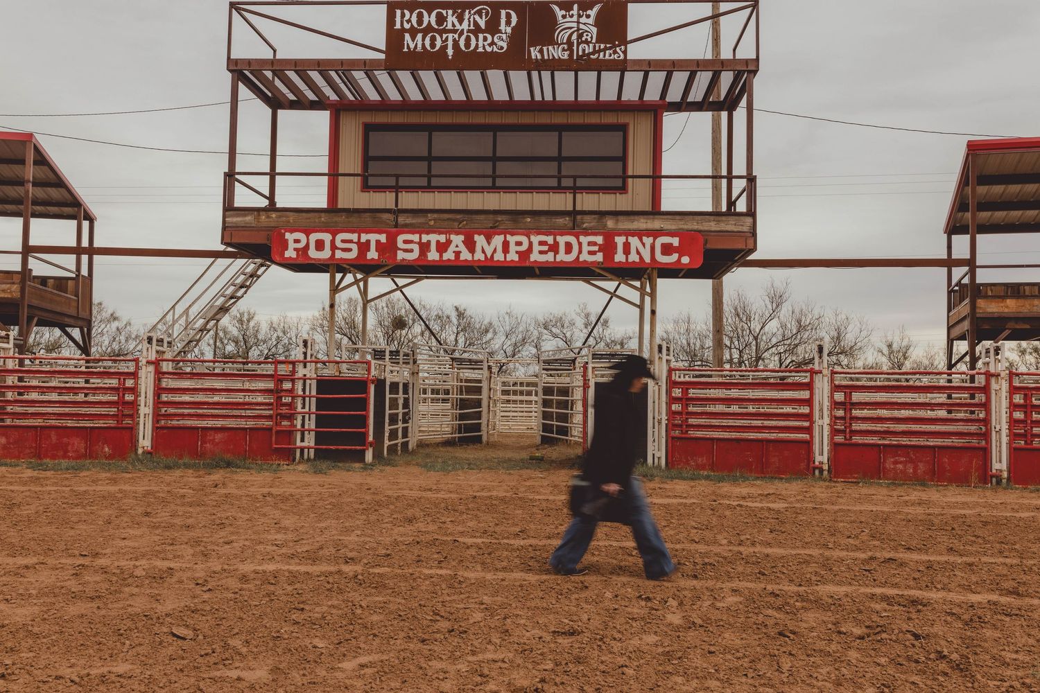 A cowboy walks through the Post Stampede Inc. rodeo arena with red fencing and grandstands on a cloudy day.