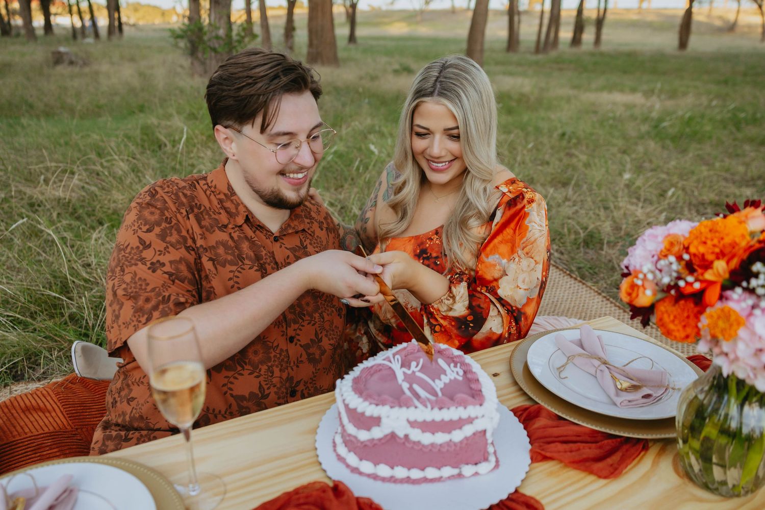 A couple in orange autumn attire shares cake at an outdoor table decorated with bright flowers during sunset.