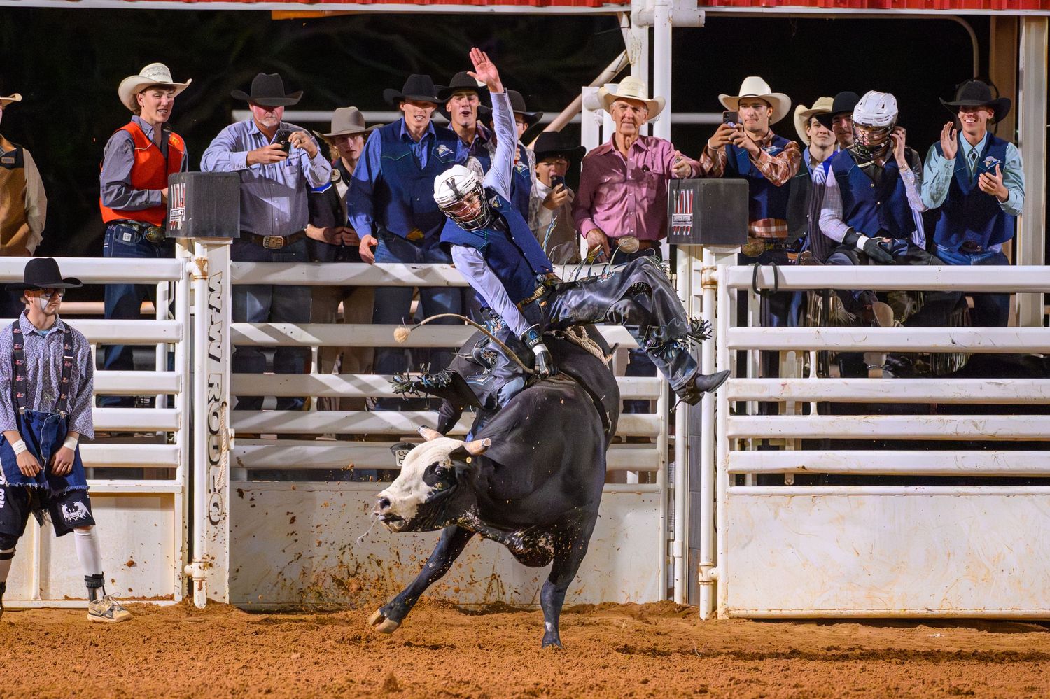 A cowboy rides a bucking bull at a rodeo arena at night, with cheering spectators and handlers watching behind rails.