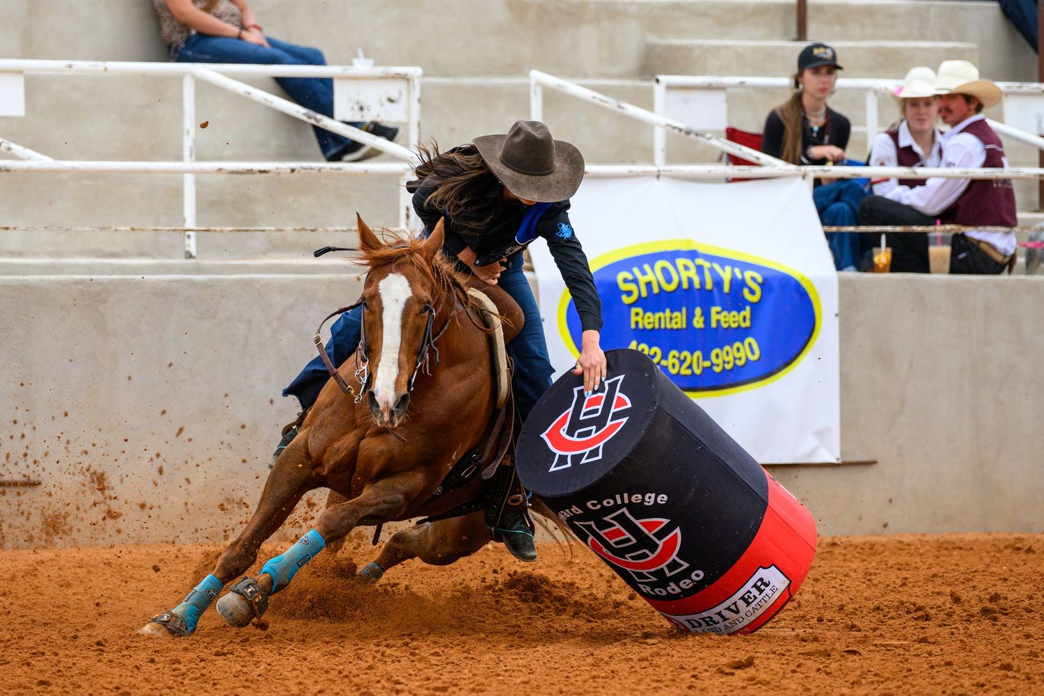 Barrel racer on chestnut horse making tight turn at rodeo arena near Shorty's Rental and Feed sign.