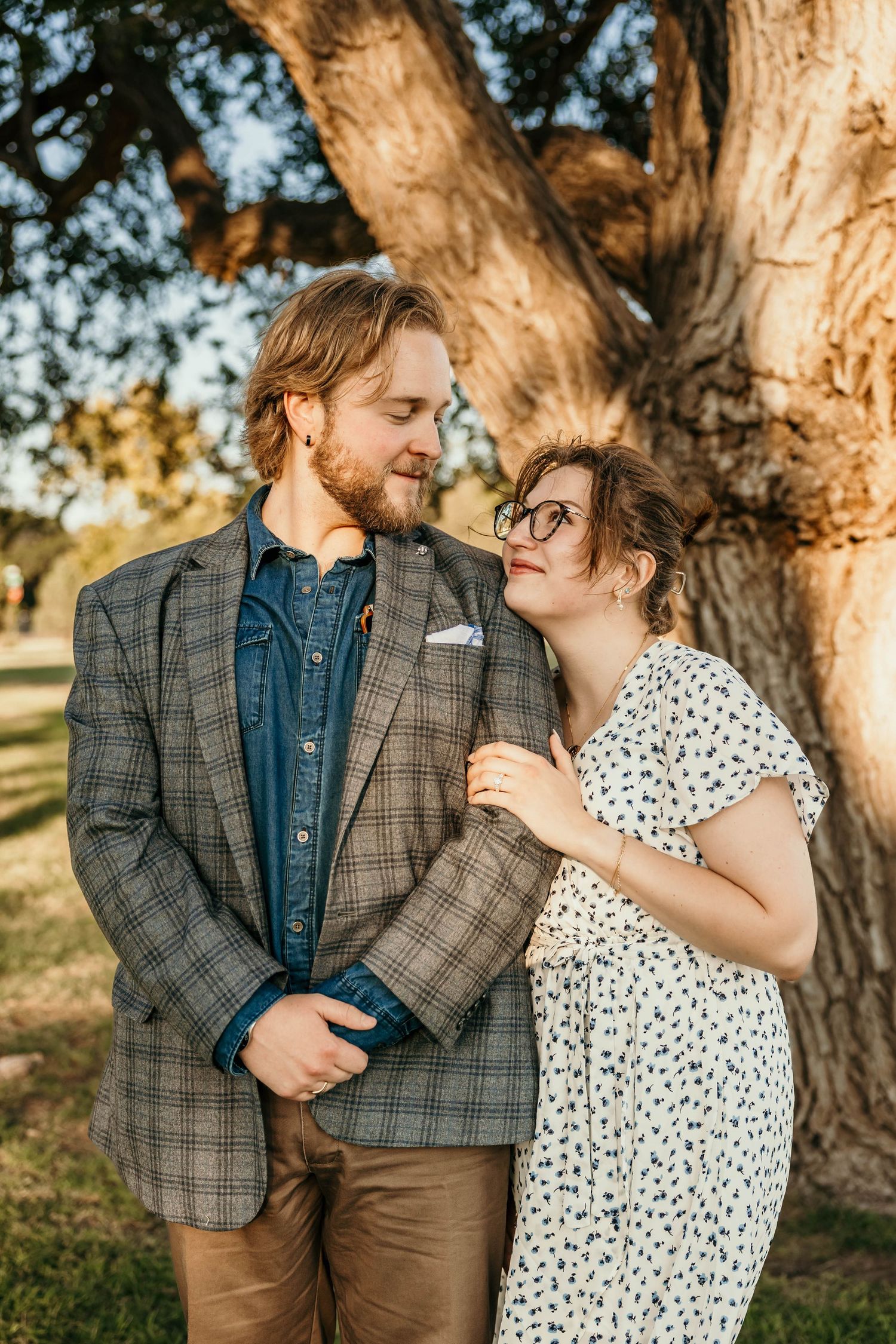A couple in polka dot dress and plaid blazer share an intimate moment under a large tree at sunset.