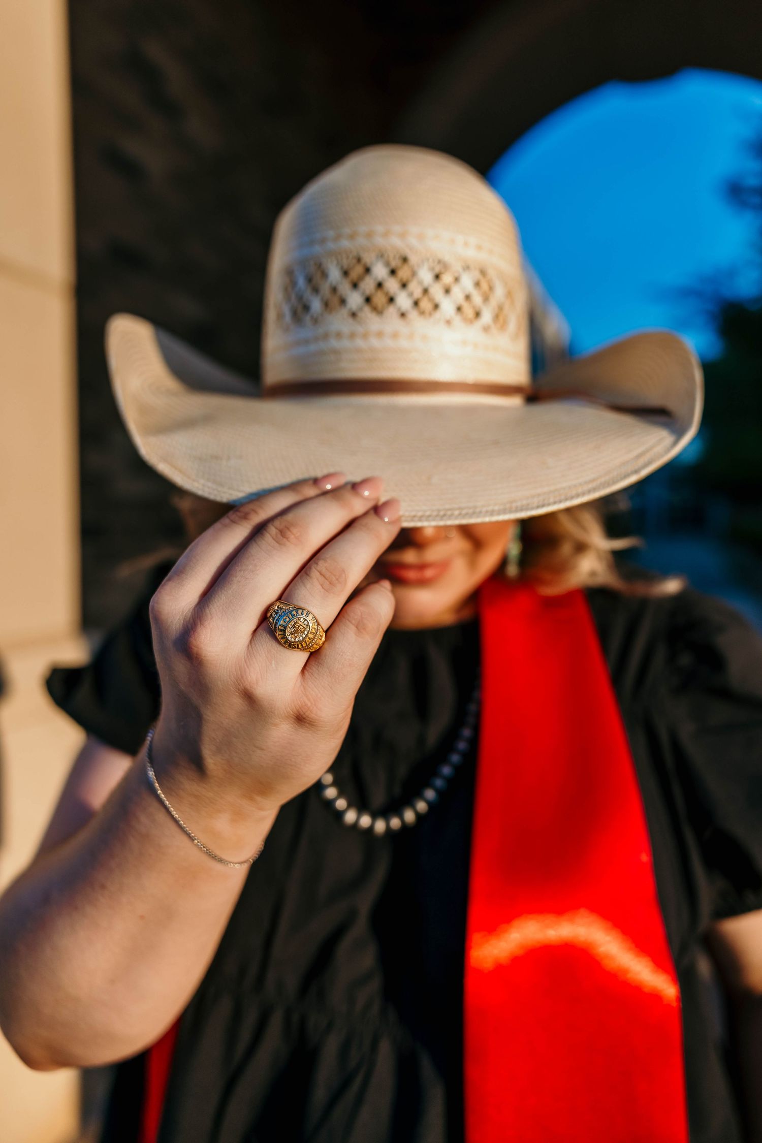 A hand tilts a light-colored cowboy hat with mesh detailing while wearing a black top and red scarf in sunlit setting.